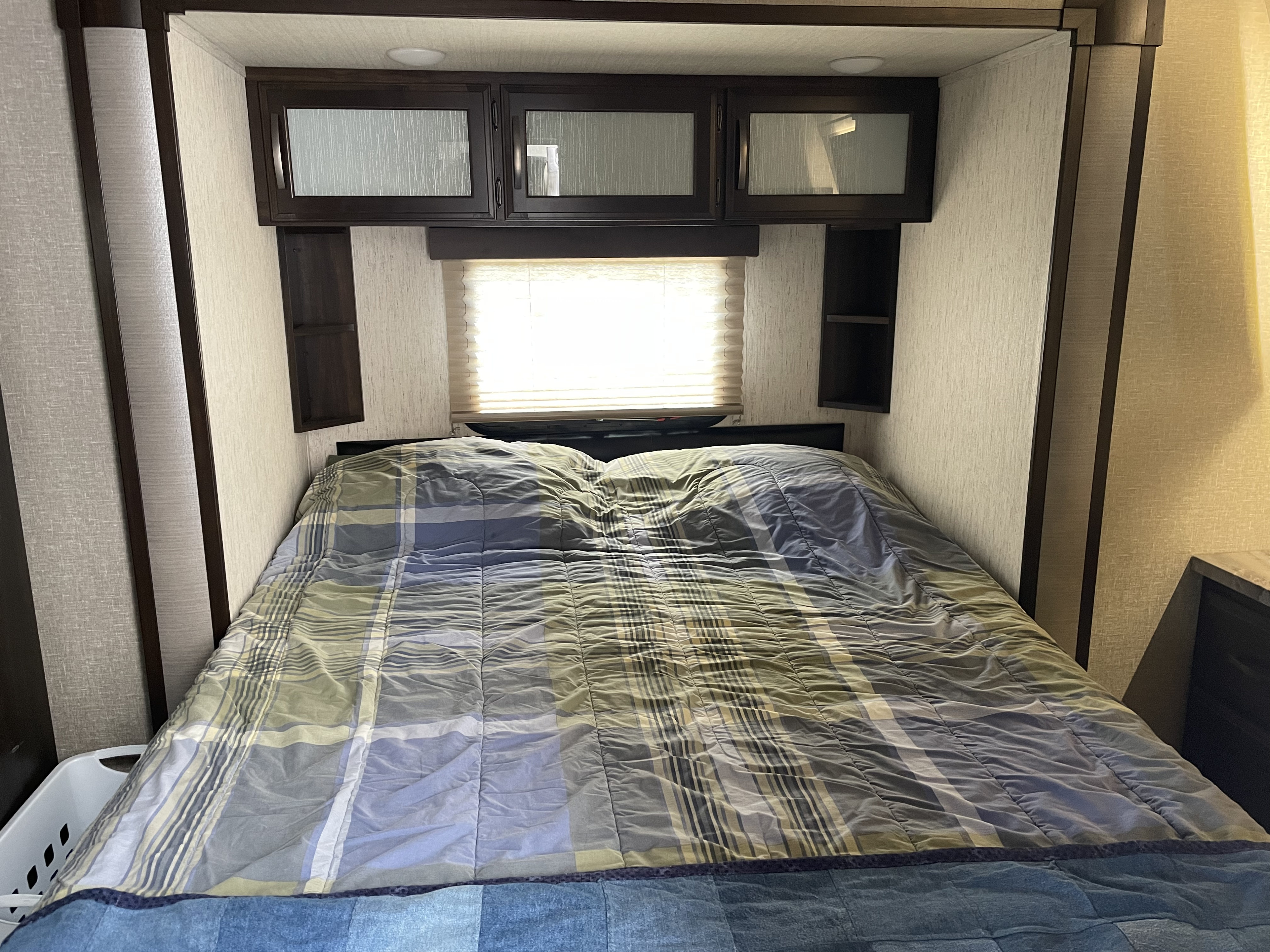 Cozy RV rental bedroom with a plaid comforter, overhead cabinets, & bright window for your travel comfort.. Grand Design Momentum 2019