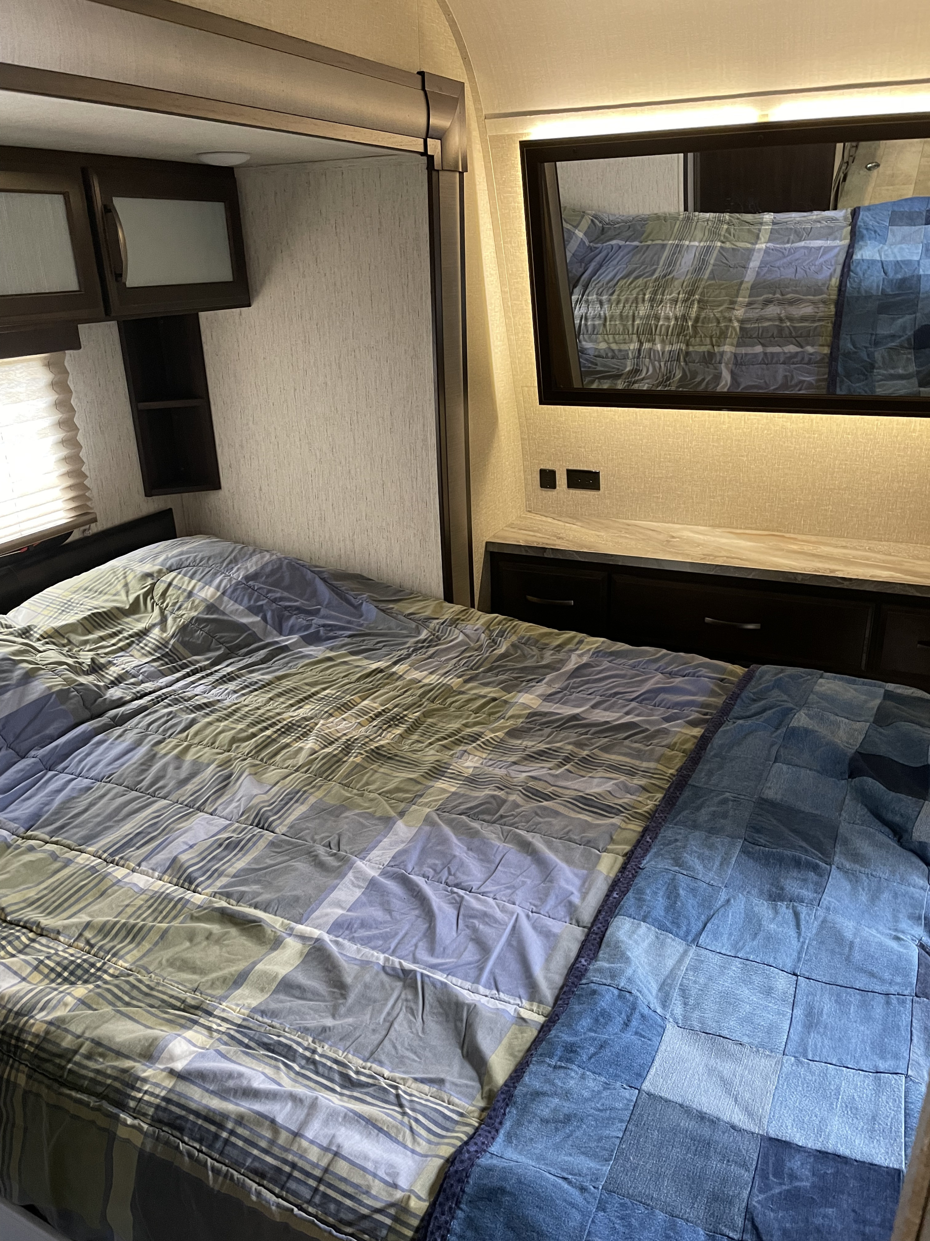 Cozy RV rental bedroom featuring a plaid bed, overhead cabinets, and a mirror/TV above a lighted countertop. Ideal for travel comfort.. Grand Design Momentum 2019
