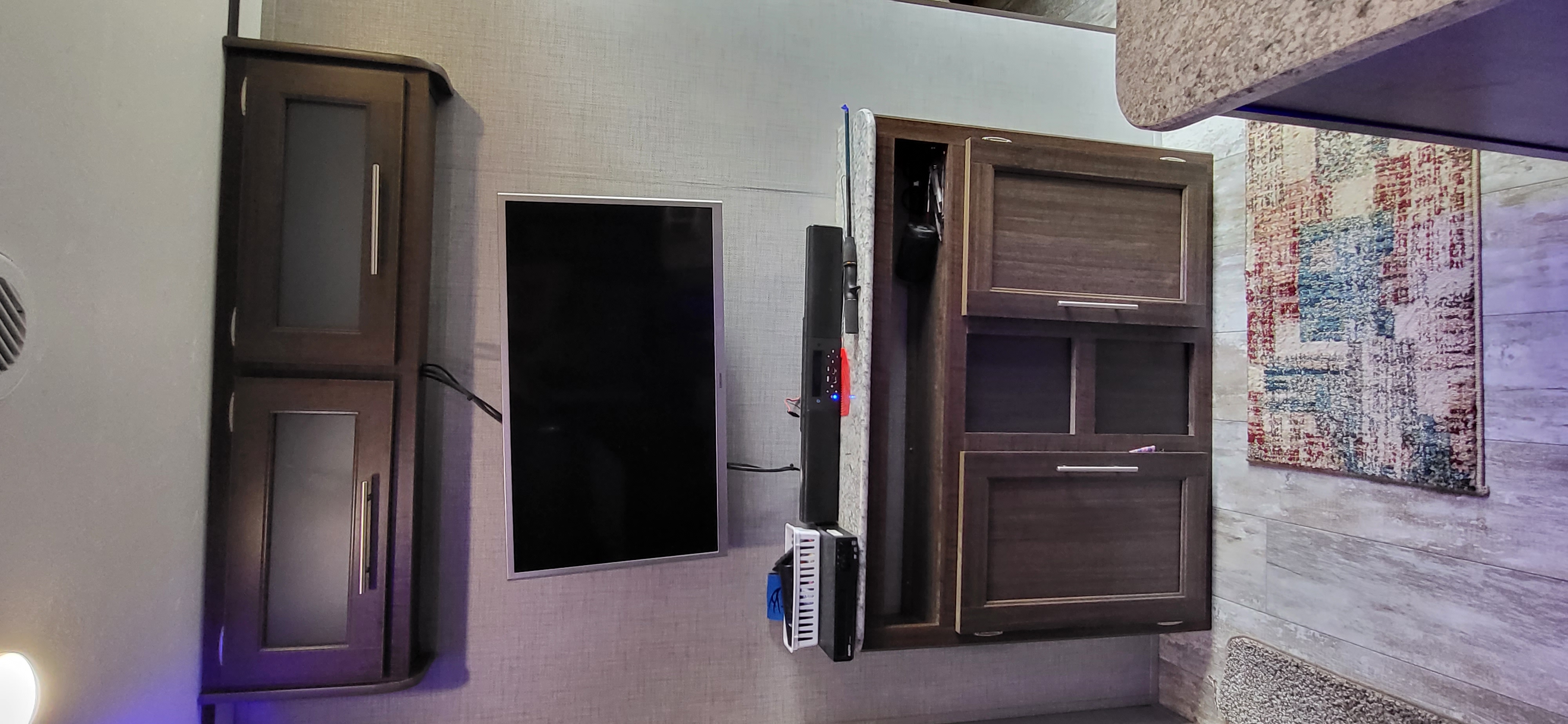 RV rental interior wall: dark cabinets, central TV, and an entertainment setup. A patterned rug hangs on a textured wall.. Crossroads RV Zinger Lite 2020