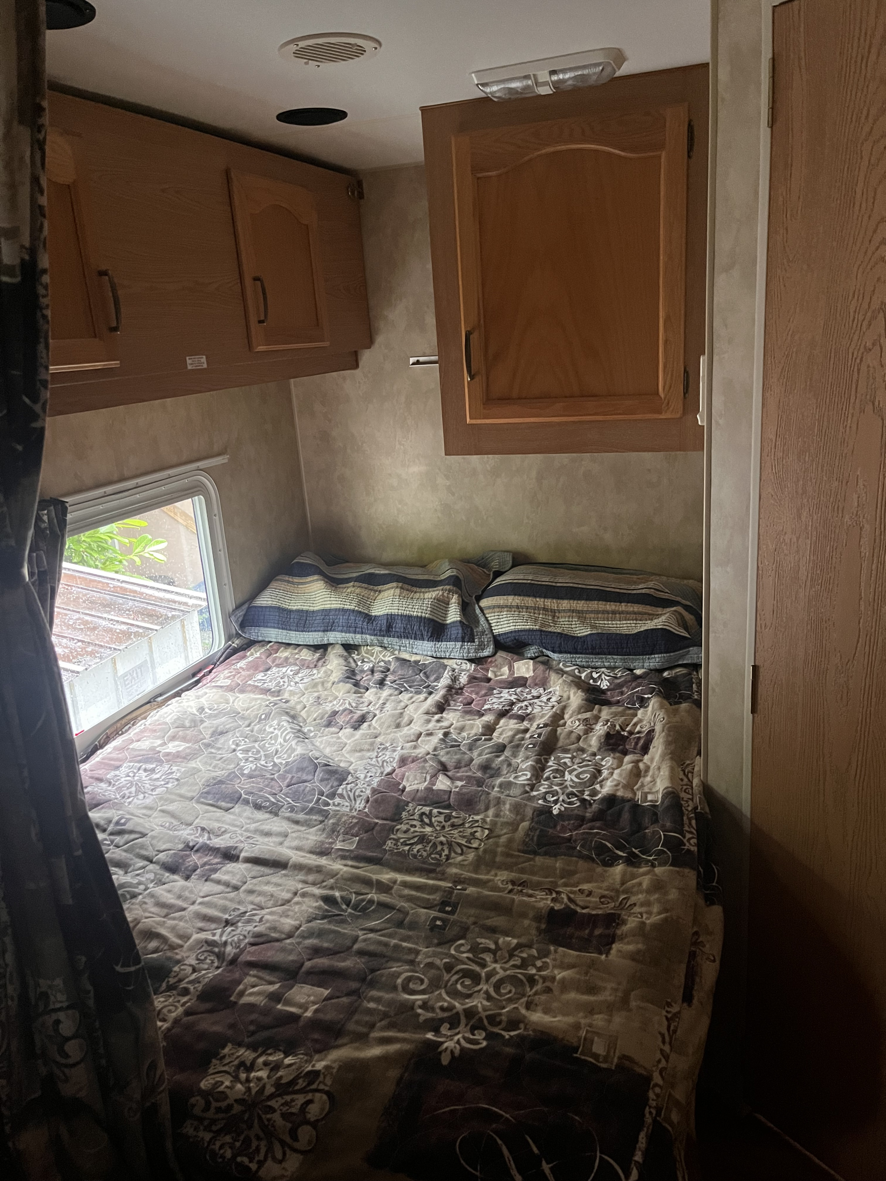 Cozy RV rental bedroom featuring a patterned bedspread, pillows, window with outdoor view, and wooden overhead cabinets.. Pioneer spirit 2007