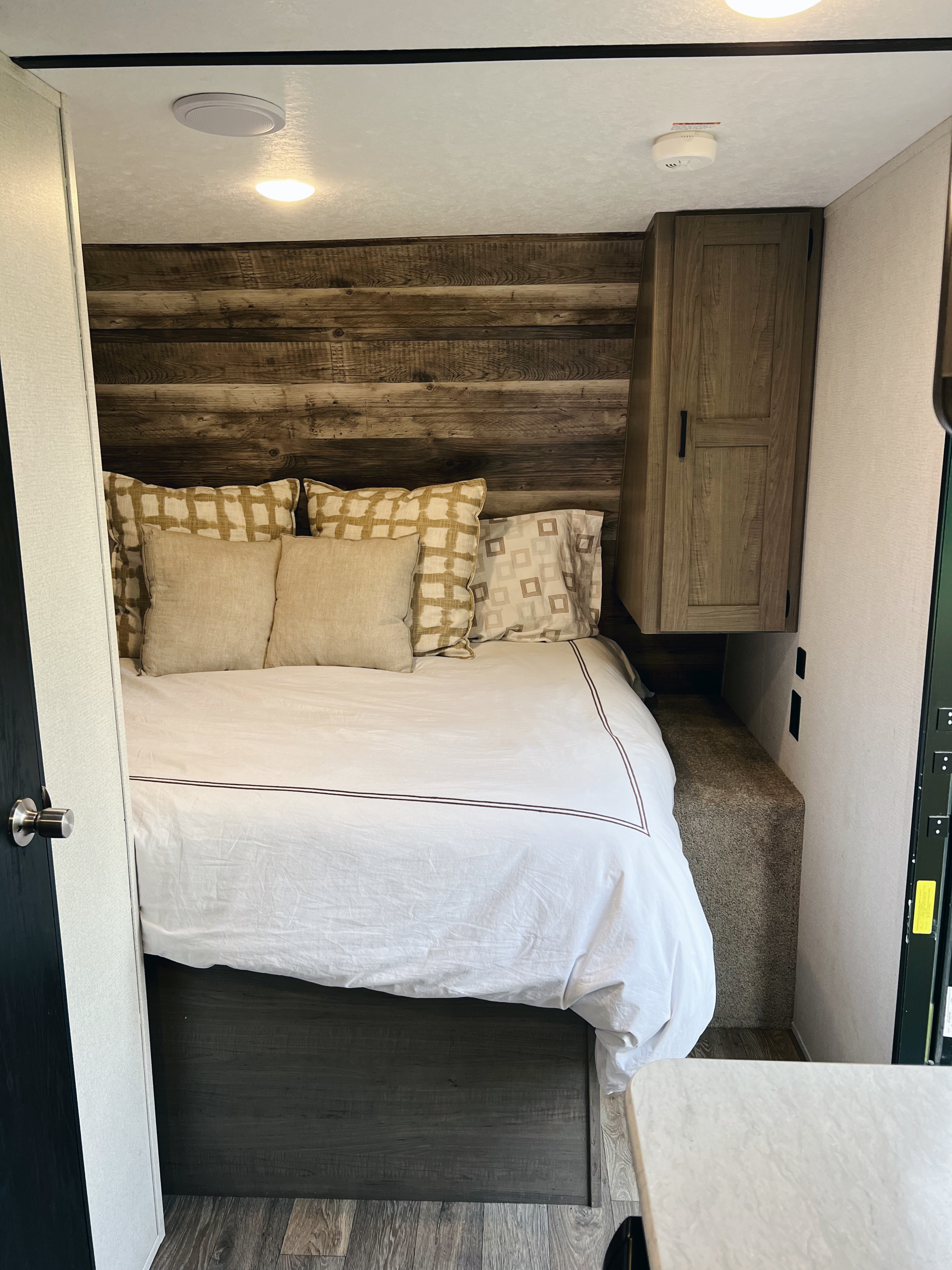 Inside an RV rental: a compact bedroom with a cozy bed, wood headboard, overhead cabinet, and bright lighting.. Keystone RV Hideout LHS Mini 2020
