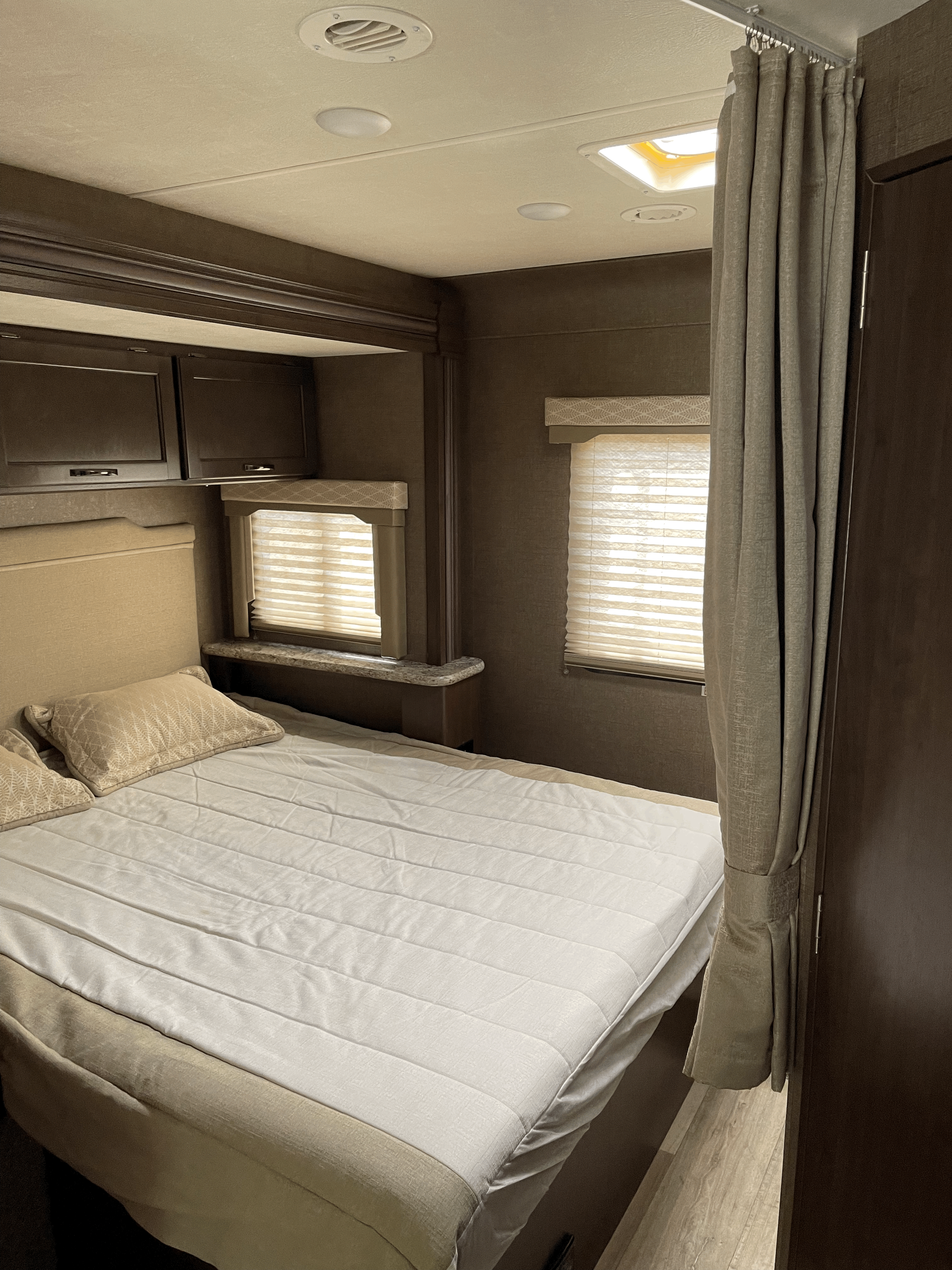 An RV rental bedroom featuring a bed with neutral bedding, two windows with blinds, overhead storage, ceiling lights/vent, and a privacy curtain.. Thor Motor Coach Freedom Elite 2019