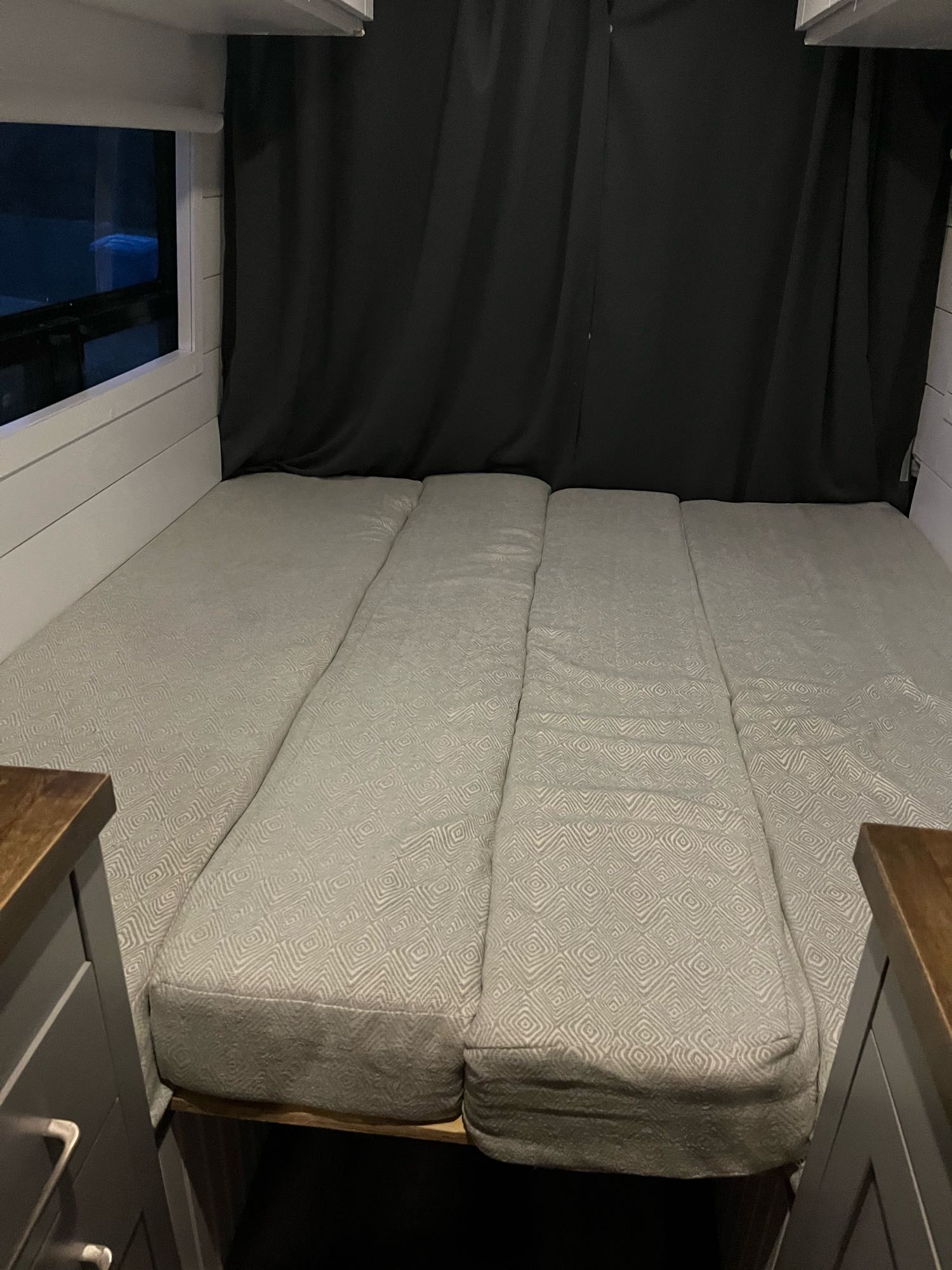 Inside an RV rental: a modular bed with light-patterned cushions, flanked by cabinets. Blackout curtains provide privacy.. Mercedes-Benz Sprinter 2013