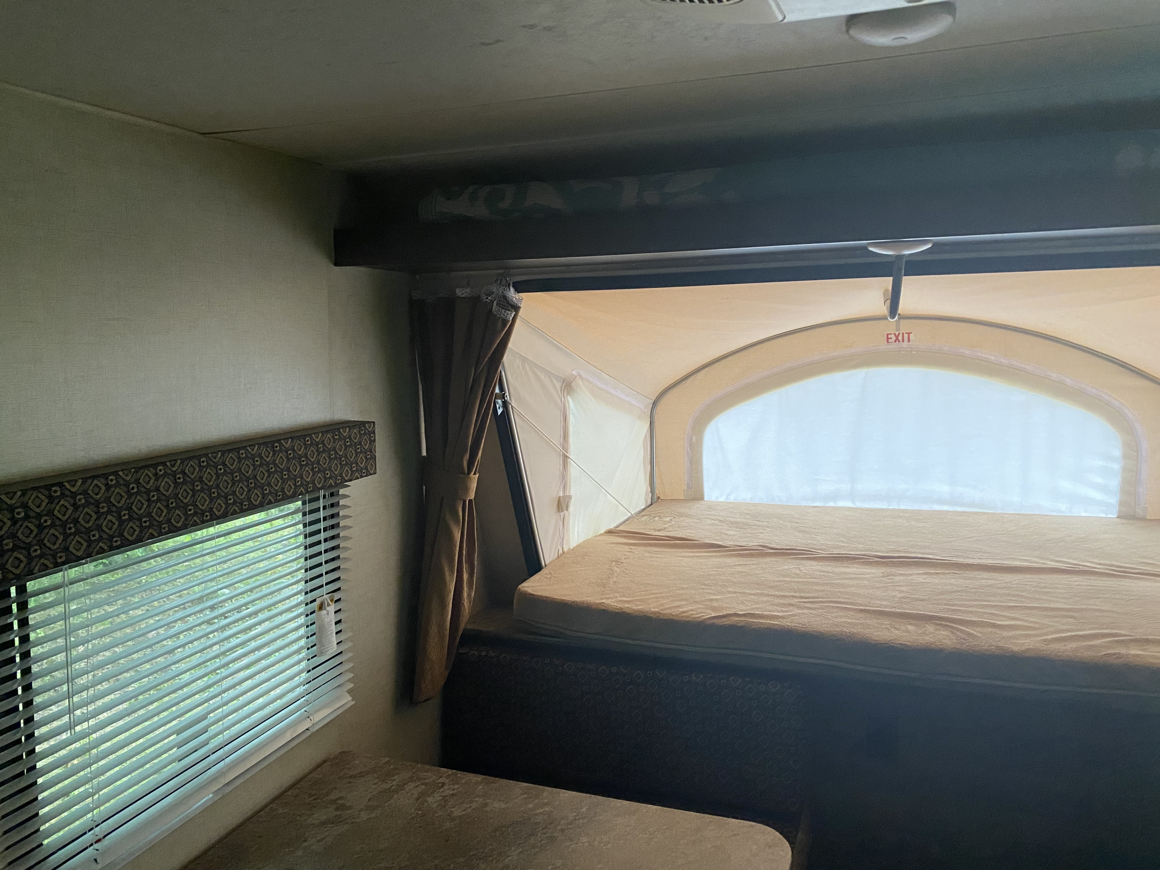 Interior of an RV rental: a cozy expandable bed with 'EXIT' sign, window with blinds, and a table.. K-Z Manufacturing Escape 2018