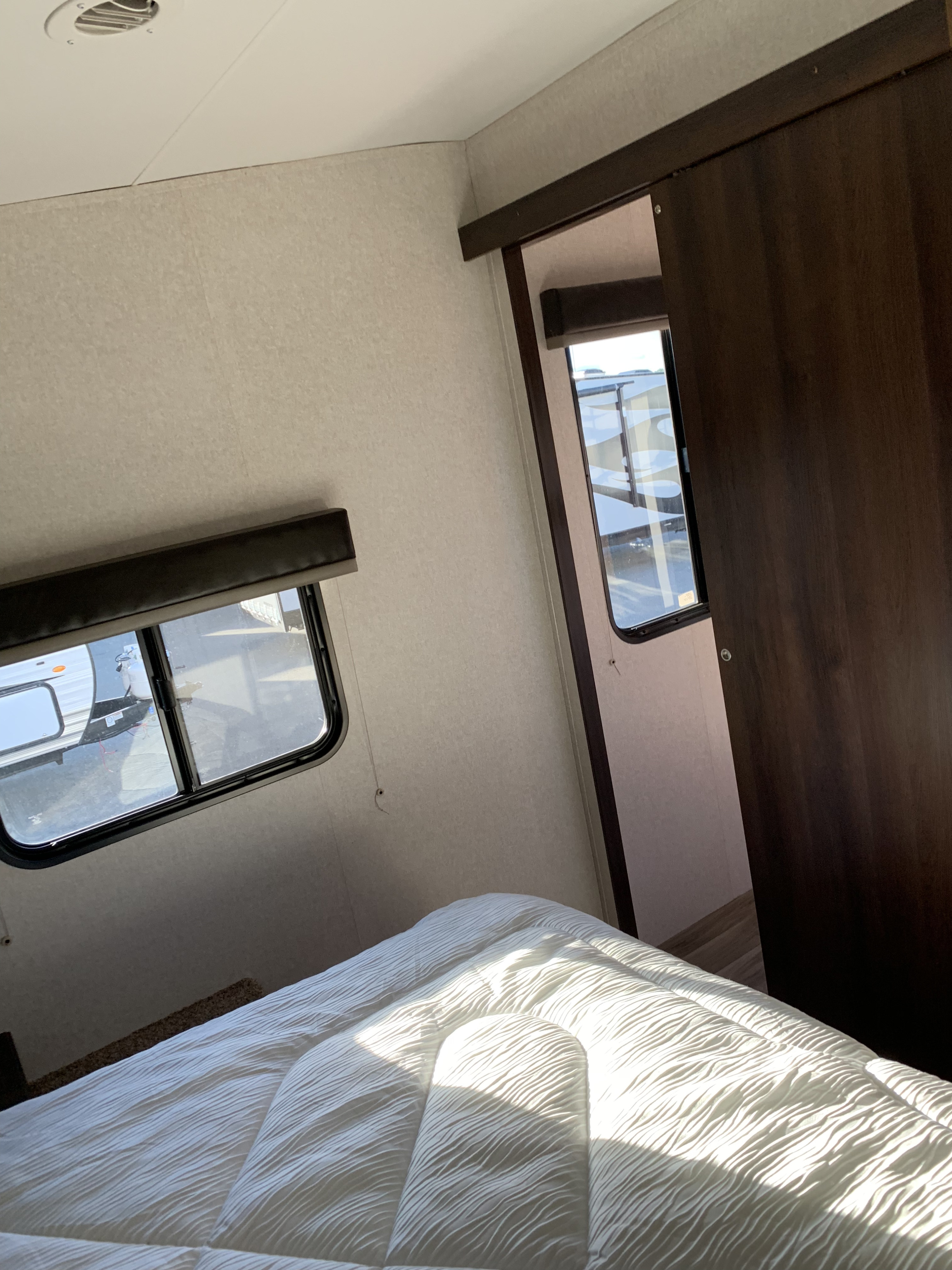 Inside an RV rental bedroom: white bedding on a bed, twin windows revealing other trailers, and a dark sliding door.. Jayco Octane 2019