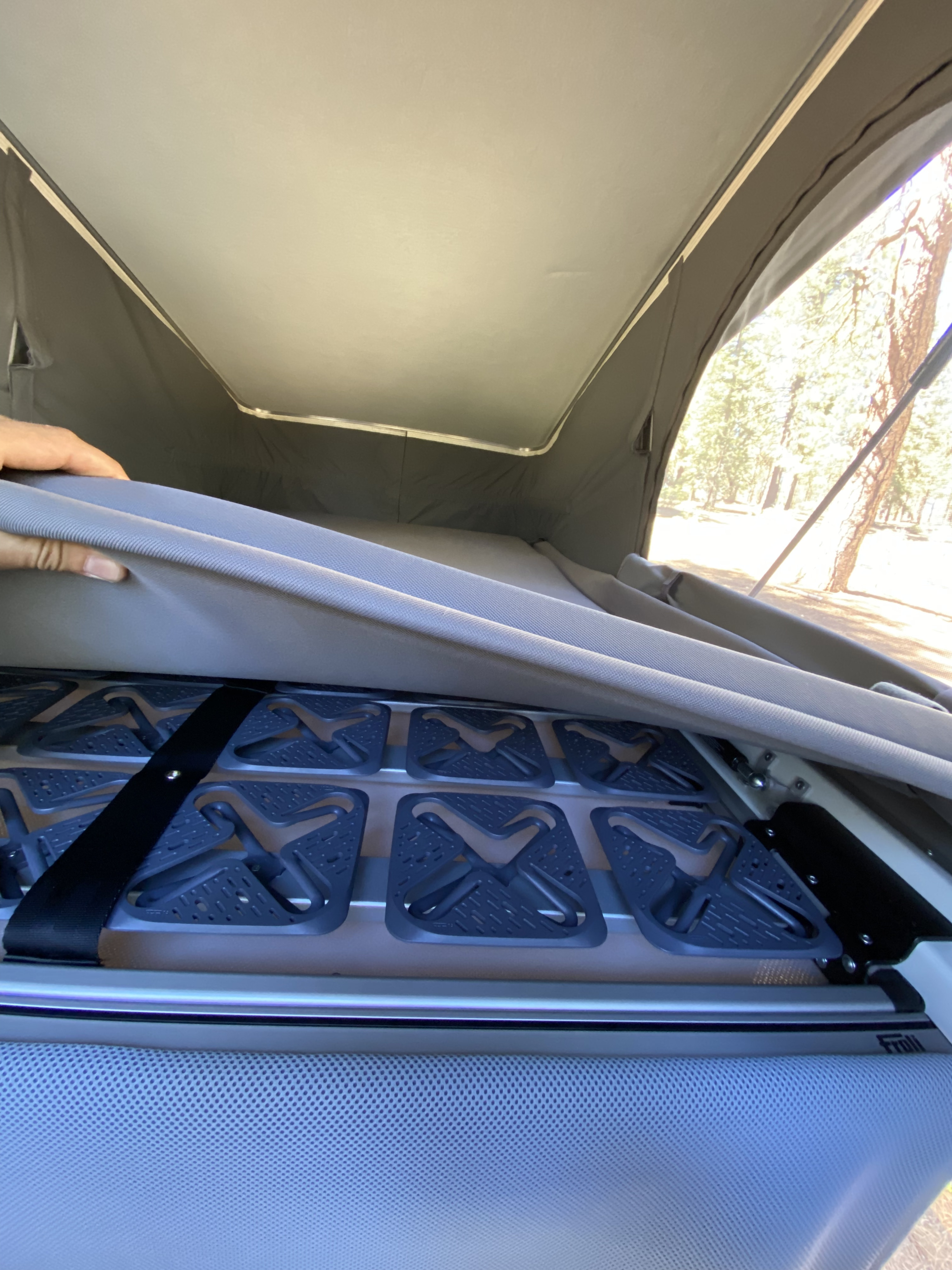 A hand lifts an RV rental mattress, revealing a Froli modular bed base for improved sleep. Pop-top tent and trees visible outside.. Mercedes-Benz Metris Weekender 2021