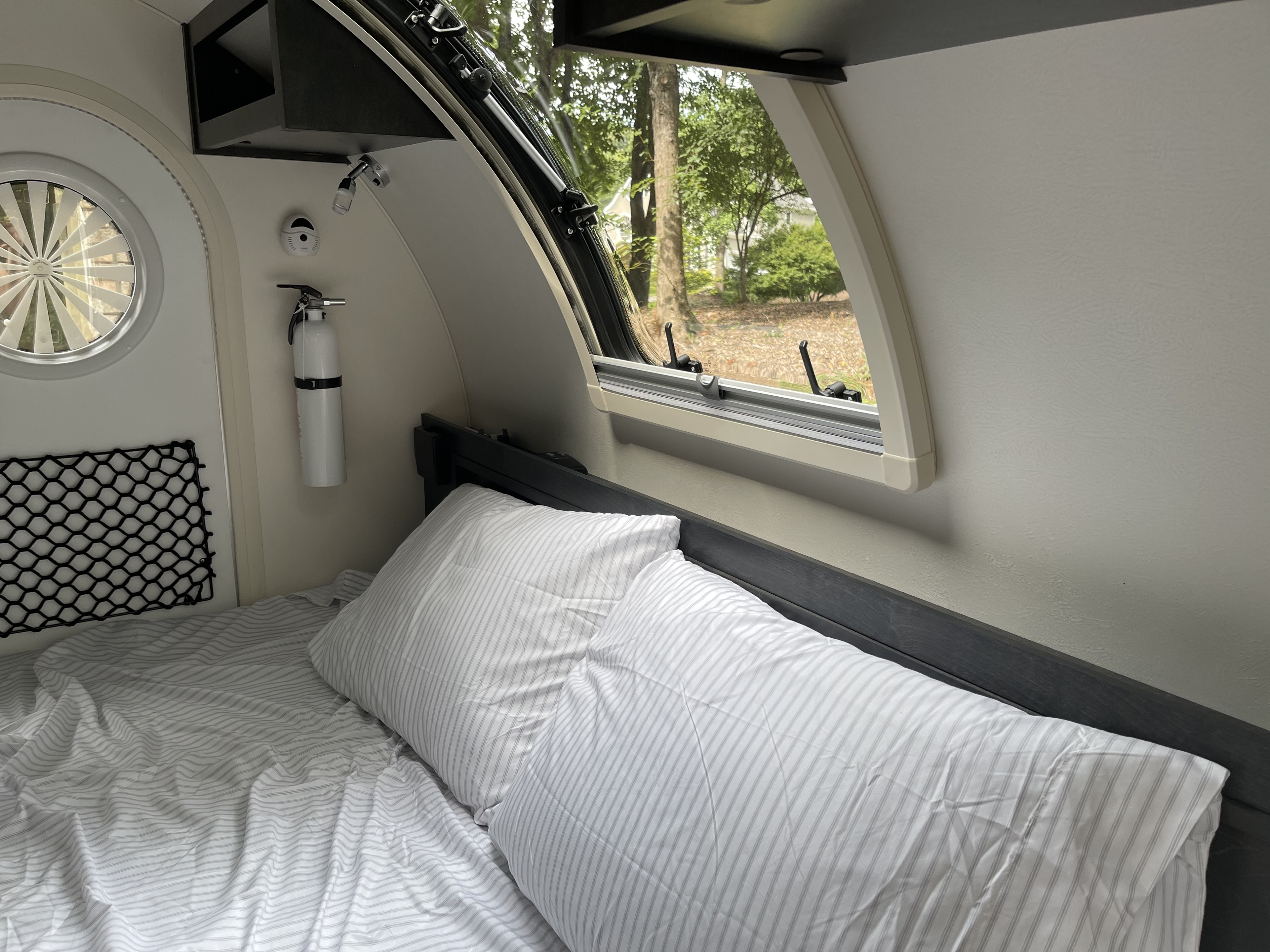 Cozy RV rental interior with a bed, window view of trees, and a fire extinguisher. Ideal for comfortable travel adventures.. nuCamp RV T@G XL 6 wide 2022