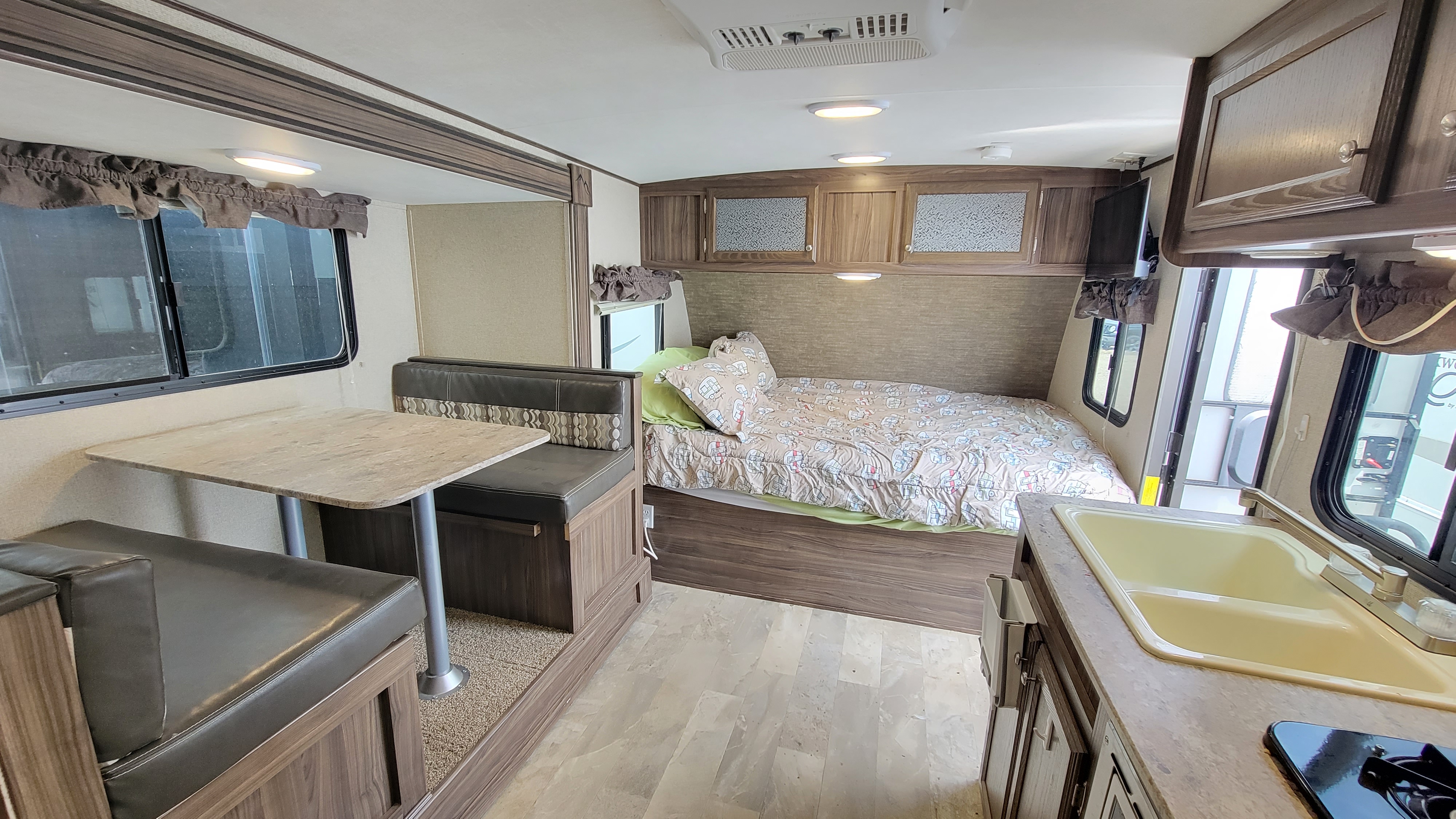 RV rental interior showing a brown dinette, bed with patterned comforter, and a kitchen area with a yellow sink.. Coachmen Apex 2018