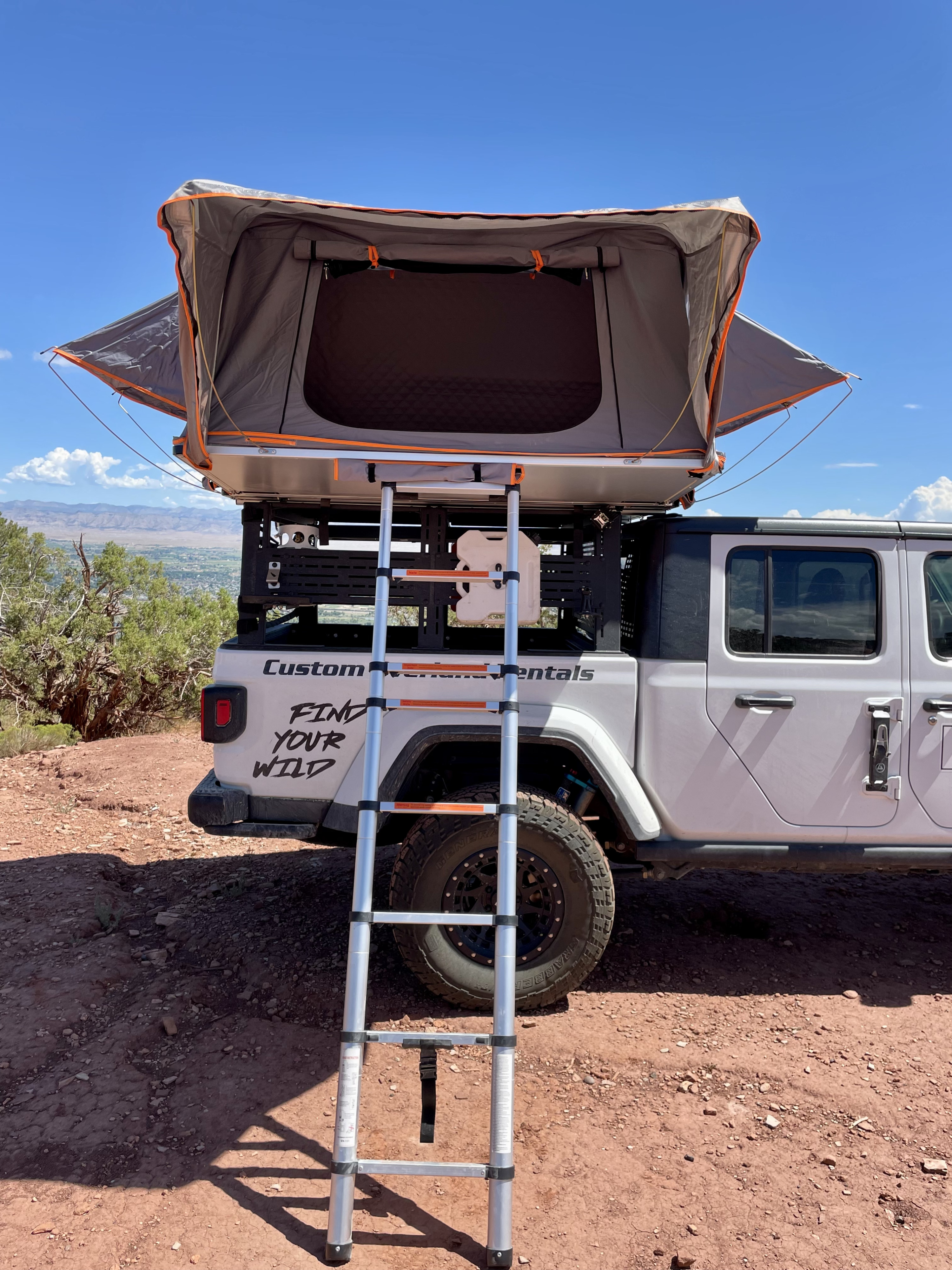 Explore with this RV rental: White Jeep Gladiator with deployed roof-top tent and ladder, ready for off-road camping adventures.. Jeep Gladiator 2021