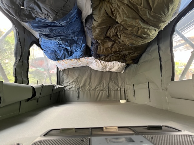 Inside an RV rental's rooftop tent, showing sleeping bags, pillows, and a window view, ready for camping trips.. ModVan CV1/M 2022