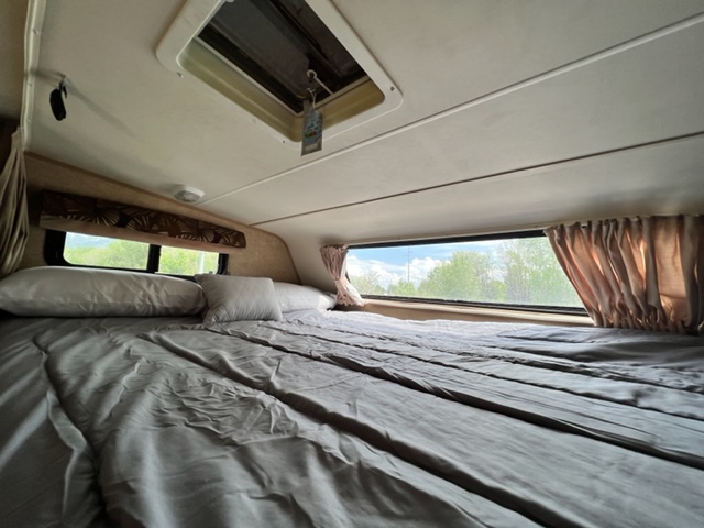 Spacious RV rental bed area with large windows, showing a comfortable, grey-covered bed and ceiling vent.. Forest River Coachmen Leprechaun 2015