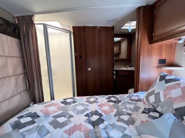 RV rental interior: cozy bedroom with patterned bed, glass shower, and adjacent bathroom with sink and toilet.. Forest River Coachmen Leprechaun 2015