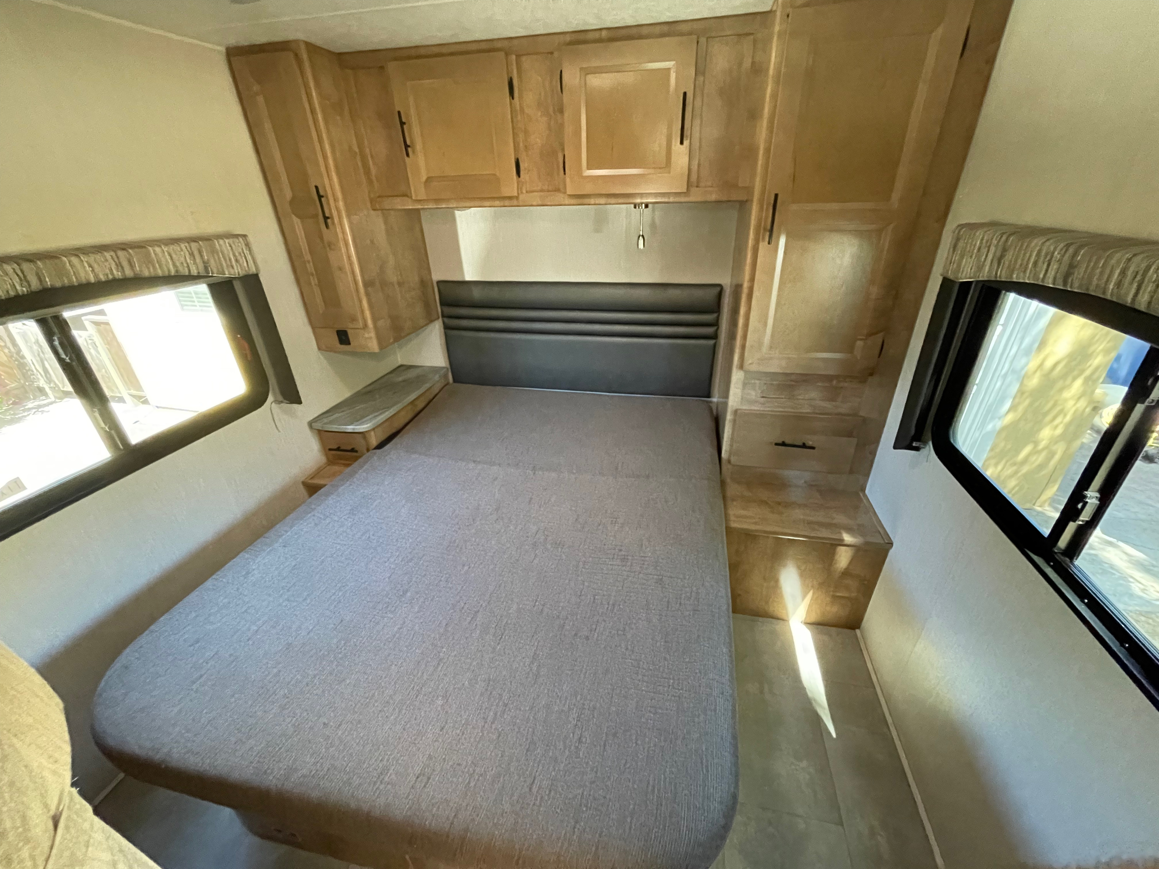 RV rental bedroom with gray bed, dark headboard, wooden overhead cabinets, and bright side windows.. Coachmen Leprechaun 2021