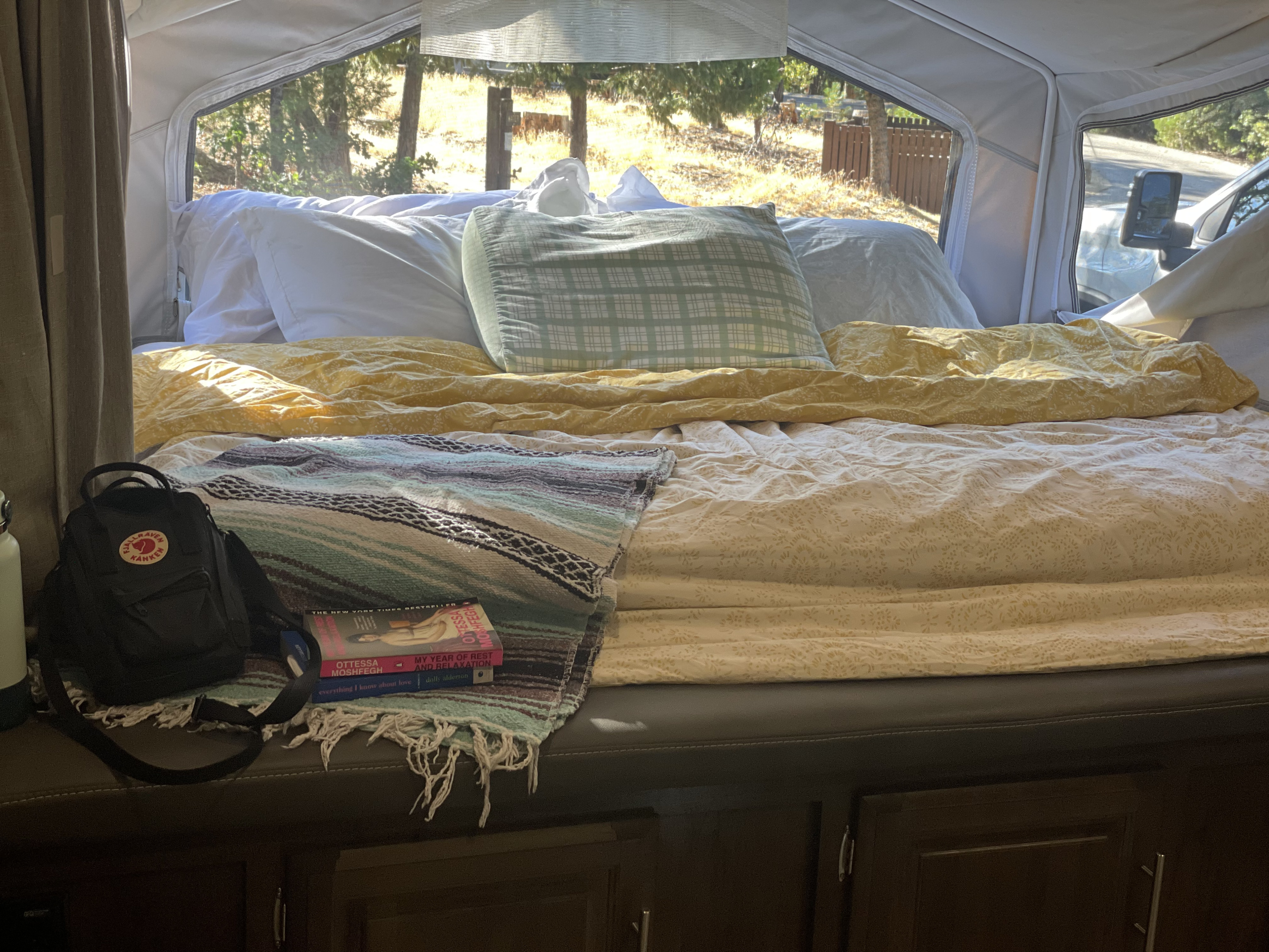 Cozy RV rental bed with pillows, patterned bedding, books, and a backpack, offering a serene nature view.. Forest River Flagstaff Shamrock 2019