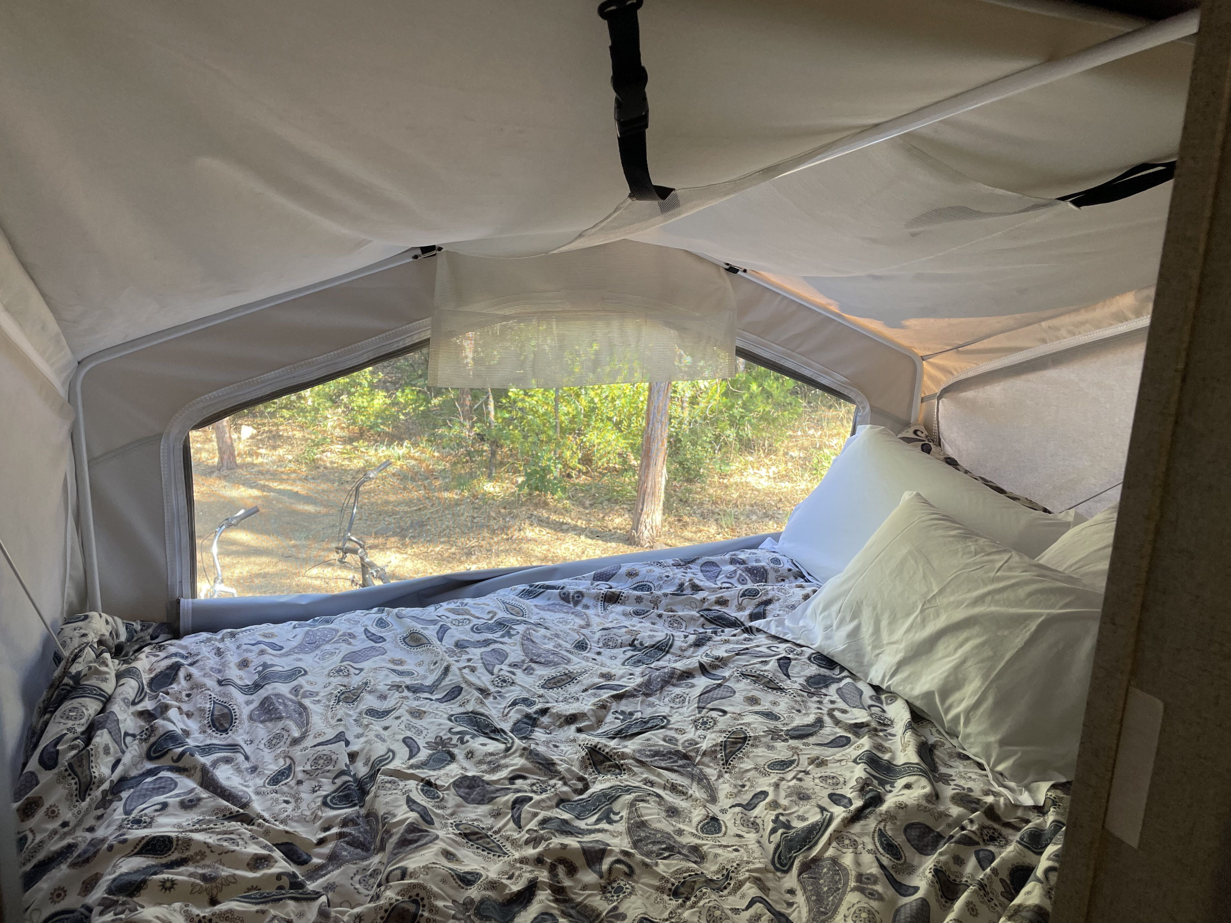 Interior of an rv rental showing a bed with paisley comforter, white pillows, and a large window with a view of trees.. Forest River Flagstaff Shamrock 2019