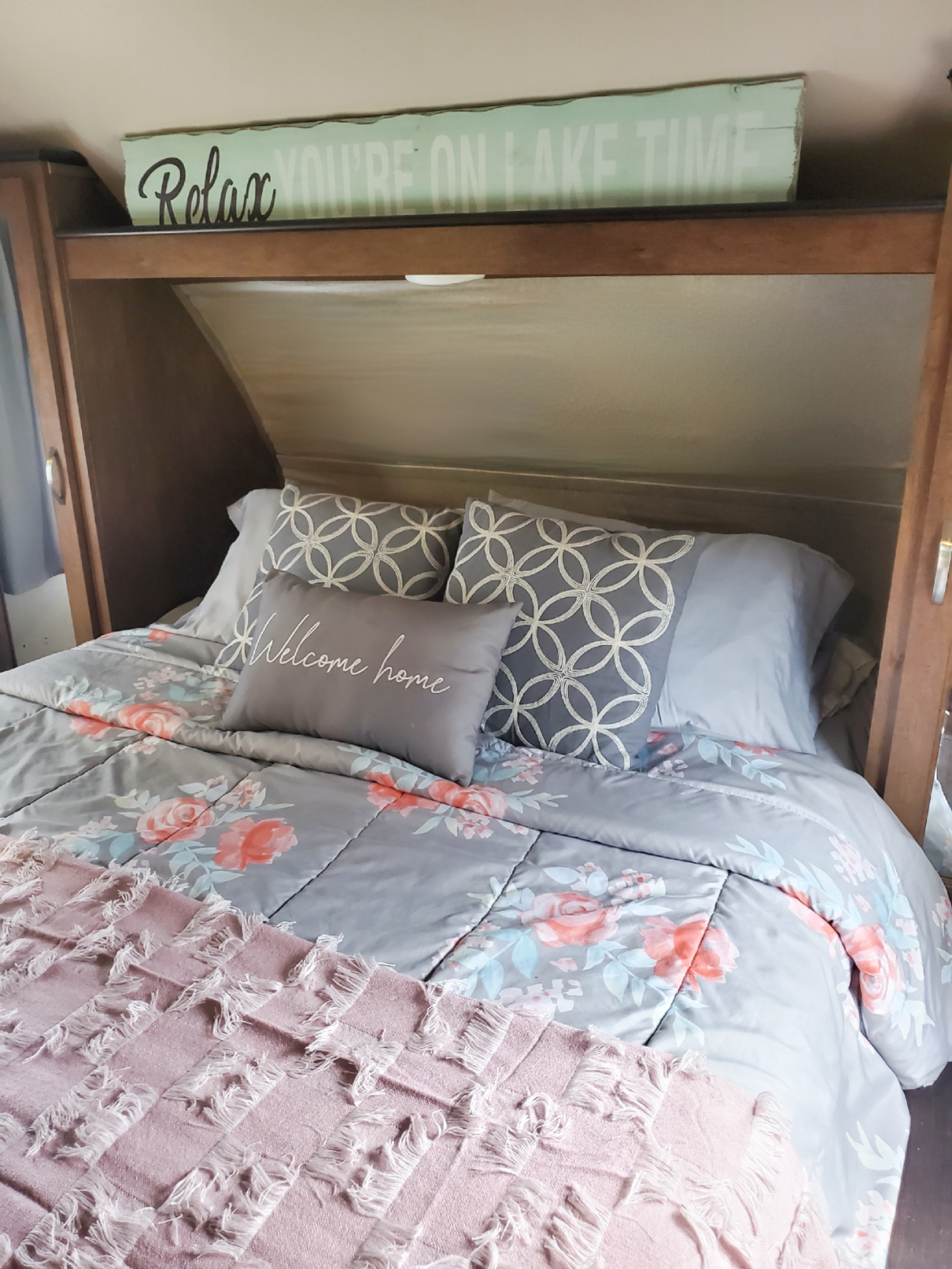 Cozy RV rental bedroom with floral bedding, "Welcome home" pillow, and "Relax YOU'RE ON LAKE TIME" sign.. Forest River Wildwood X-Lite 2017