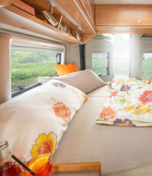 Cozy RV rental interior: a comfy bed with floral bedding, scenic window views of nature, and a refreshing drink.. Citroen Jumper 2022