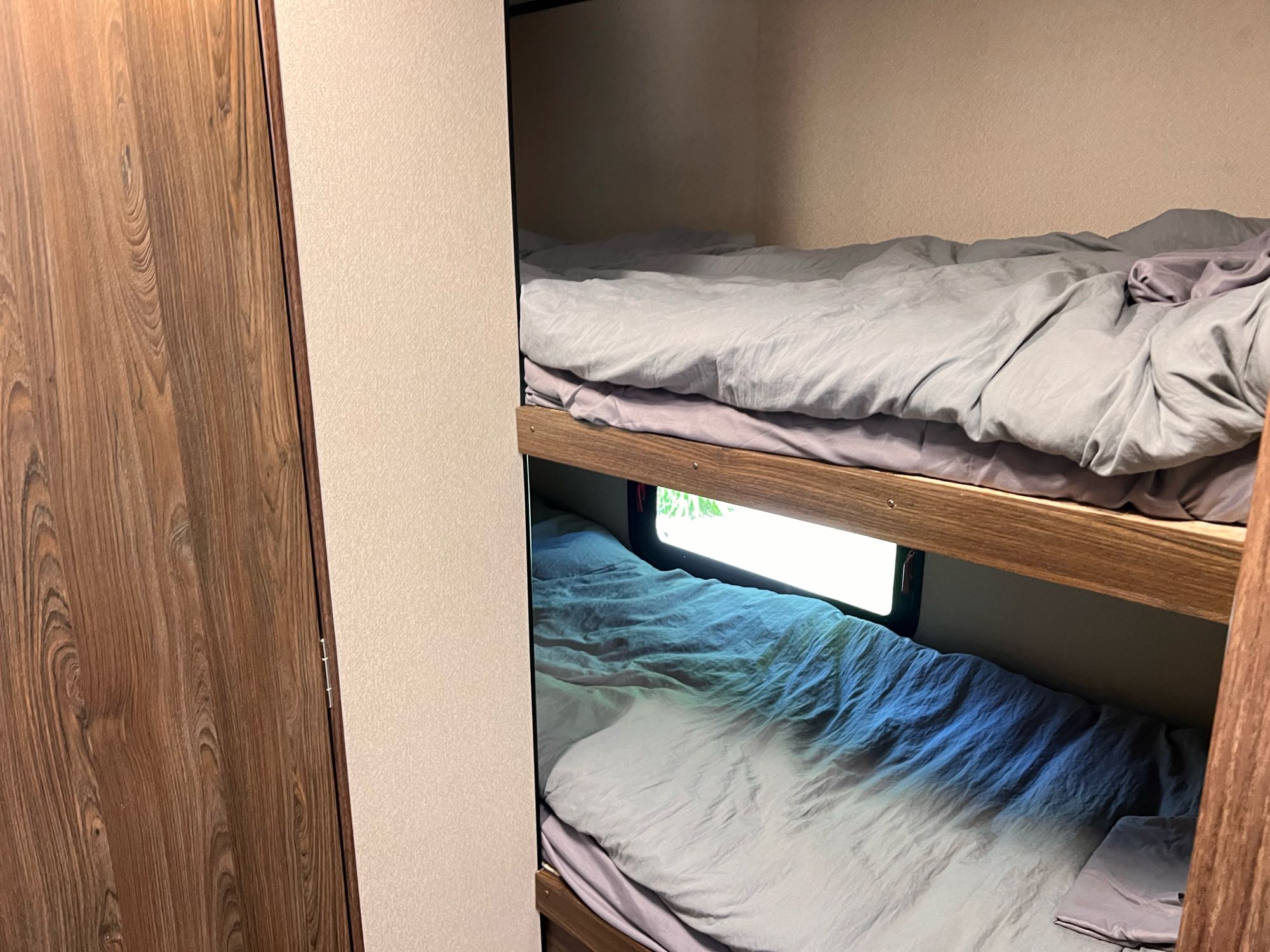 Inside an RV rental: two bunk beds with light bedding. A wood cabinet is on the left, and a window offers a view from the lower bunk.. Riverside Travel Trailer Dream 2018