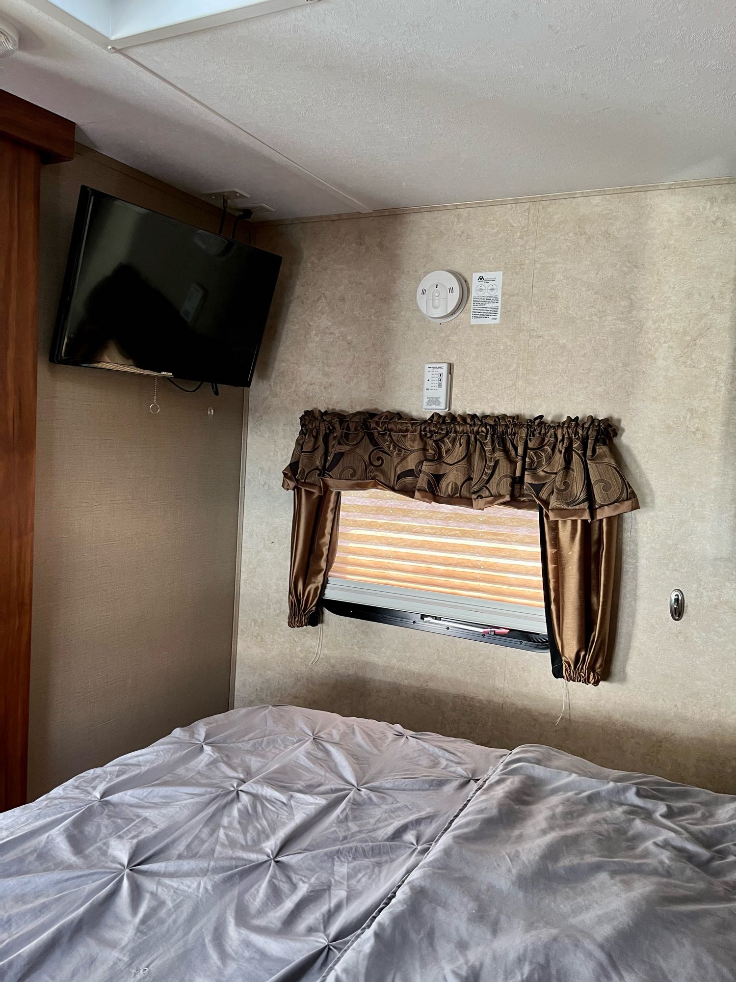 RV rental bedroom features a mounted TV, window with brown curtains, and a comfortable gray bed, ideal for cozy travels.. Keystone 29RBKWE 2015