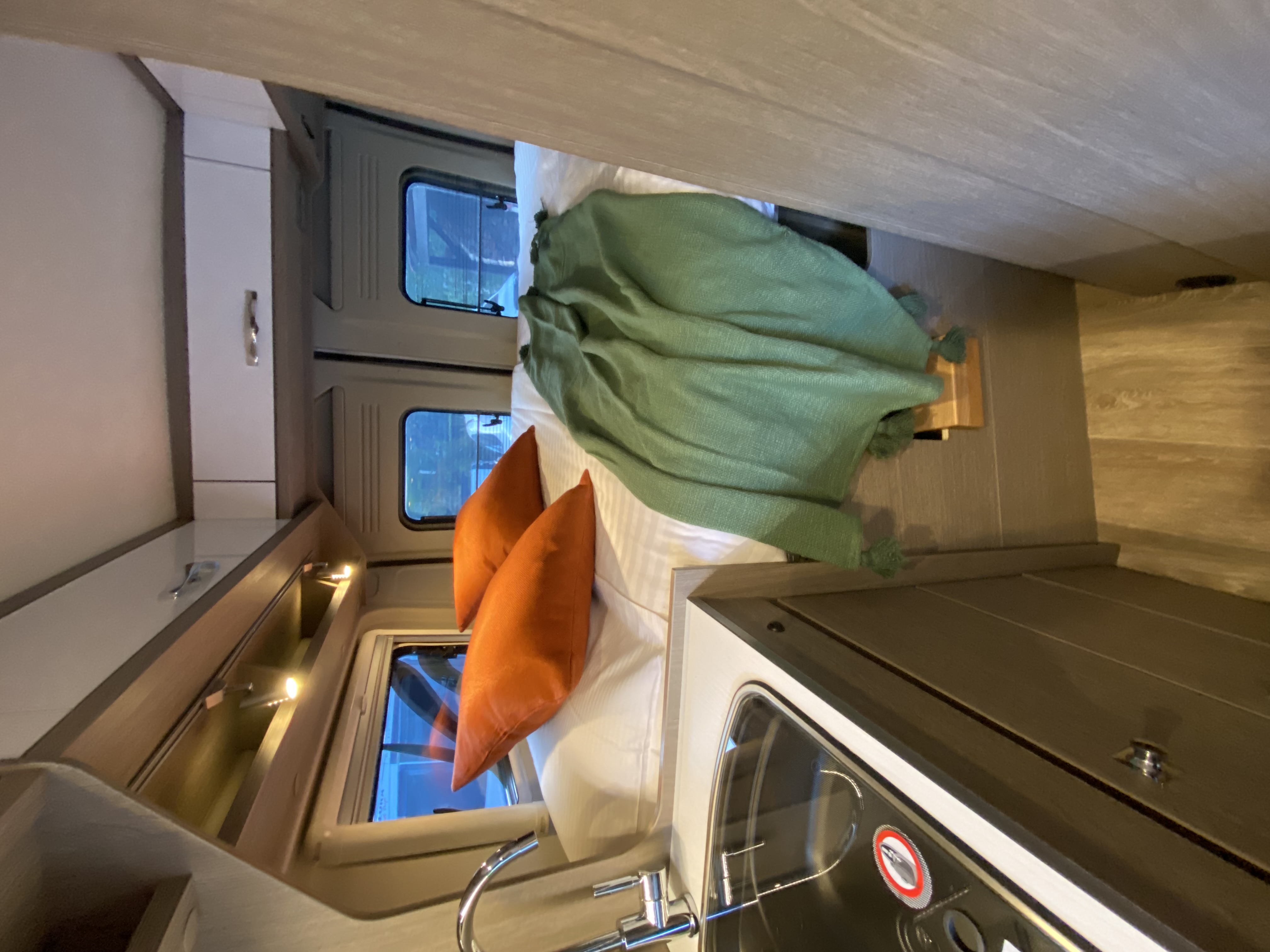 View of a compact RV rental interior, showcasing the bed with green blanket & orange pillows, windows, and kitchenette.. Citroen Jumper 2022