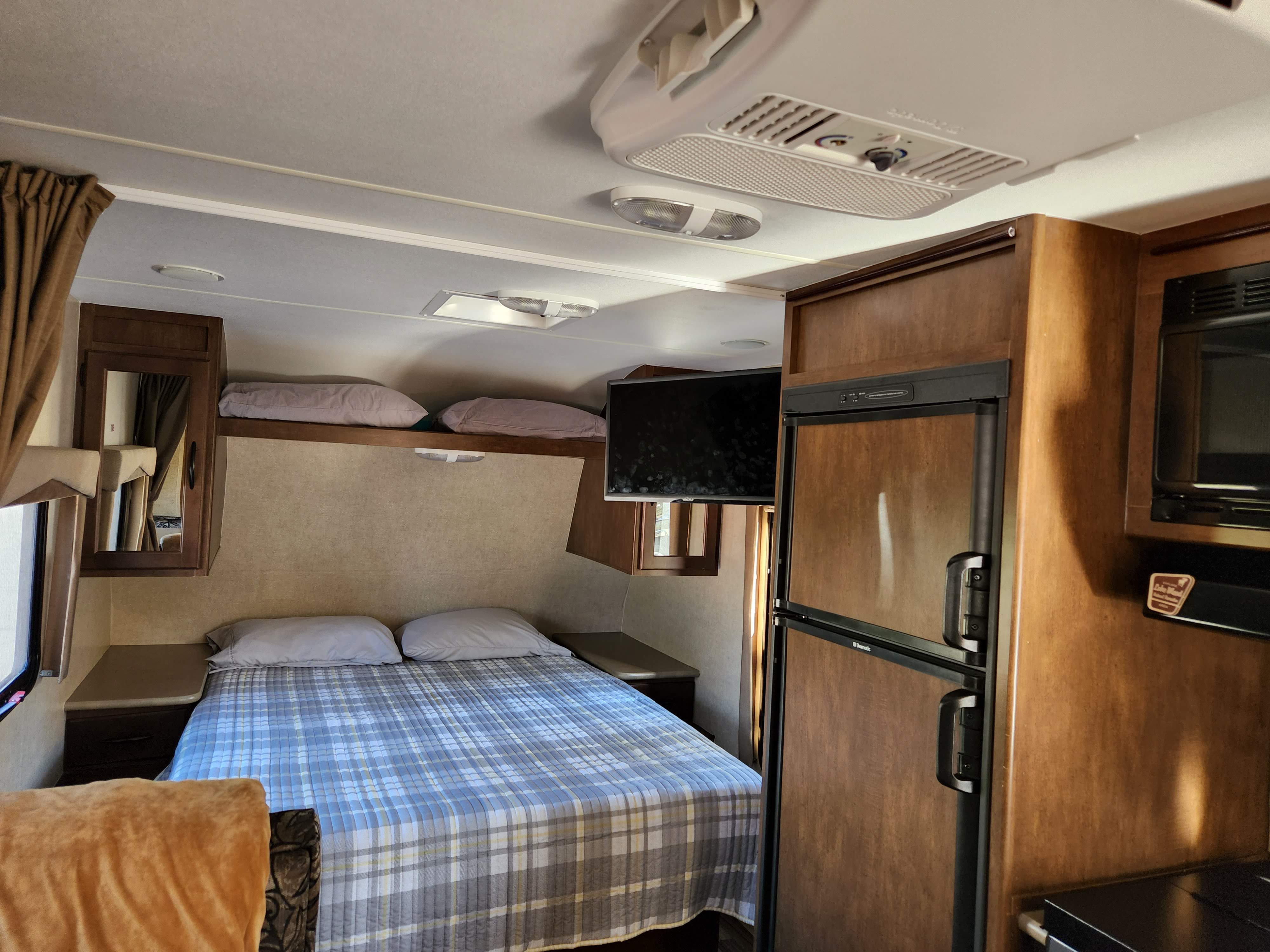 Inside this RV rental, find a compact bedroom with plaid bedding, overhead storage, TV, and adjacent kitchen area with fridge and microwave.. Pacific Coachworks Tango Ultralite 2016
