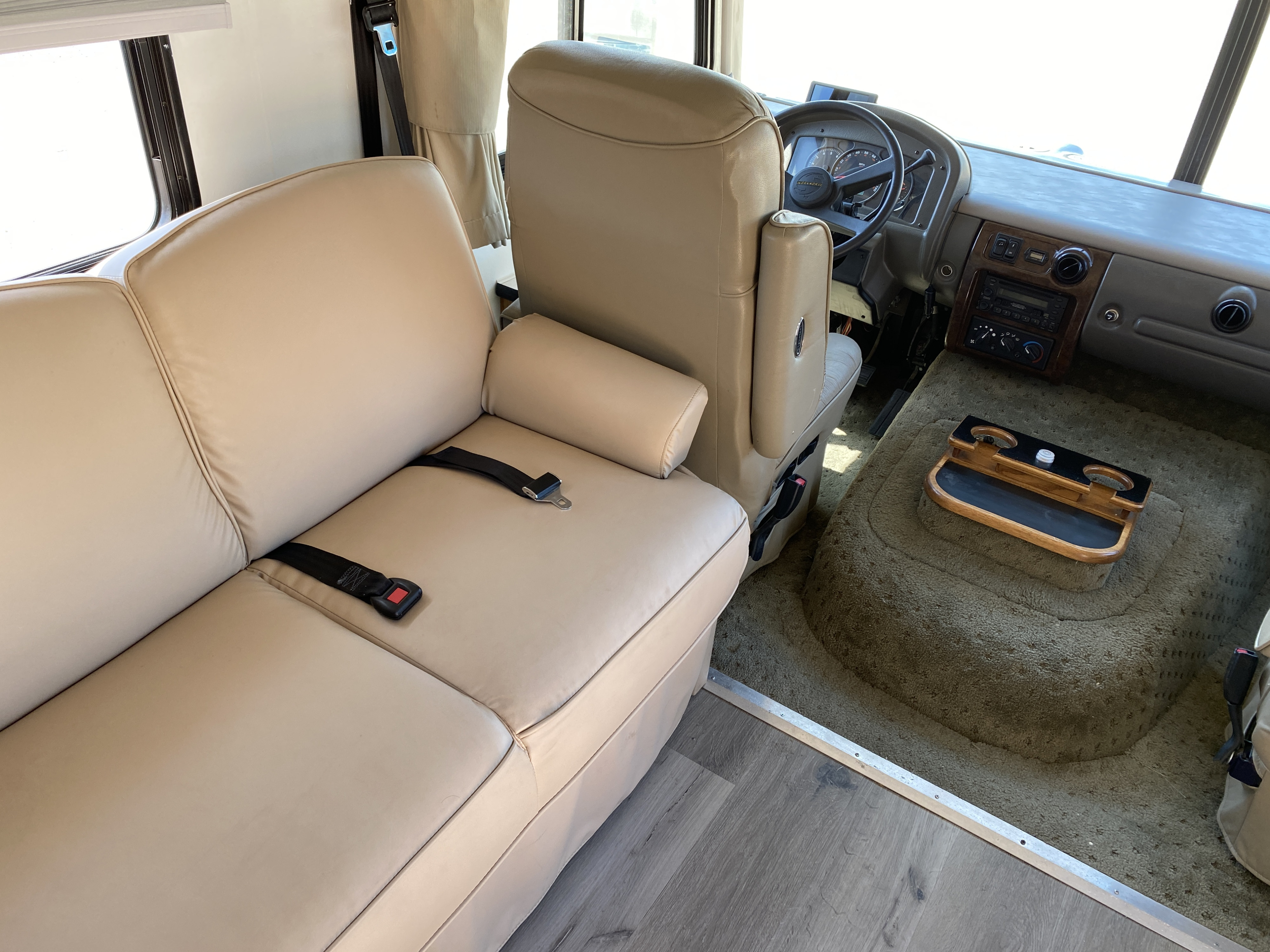 RV rental interior: Light beige sofa with seatbelts, driver's dashboard, and mixed wood/carpet flooring. Ready for travel.. Flair-Fleetwood Flair-Fleetwood Motorhome 2004