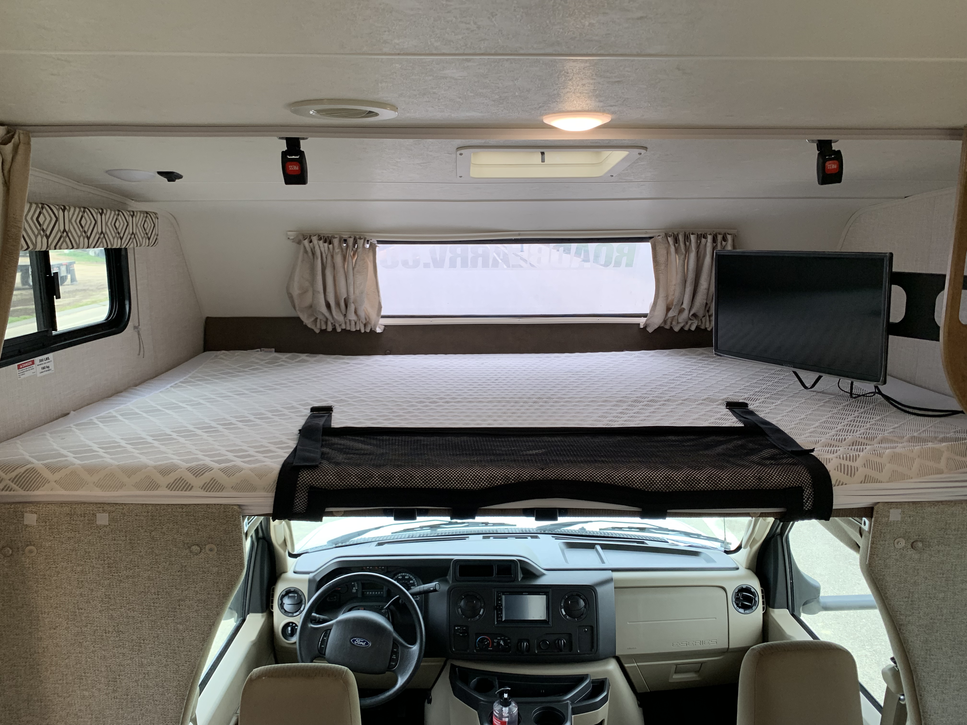Interior of an RV rental showing the cab-over bunk bed with mattress, safety net, TV, and the driver's cockpit with Ford steering wheel.. Forest River Coachmen Leprechaun 2020