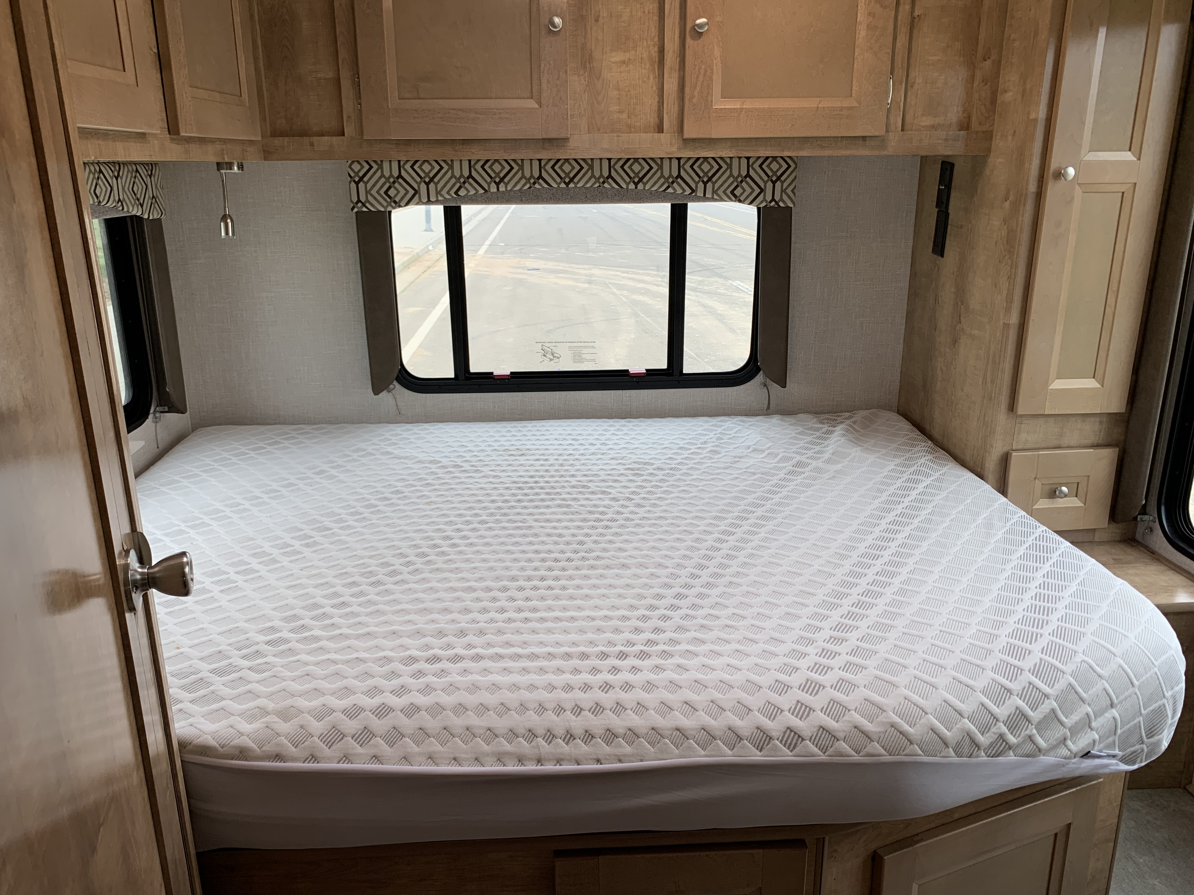 RV rental bedroom: cozy space with large bed, overhead cabinets, window view, and light fixture.. Forest River Coachmen Leprechaun 2020