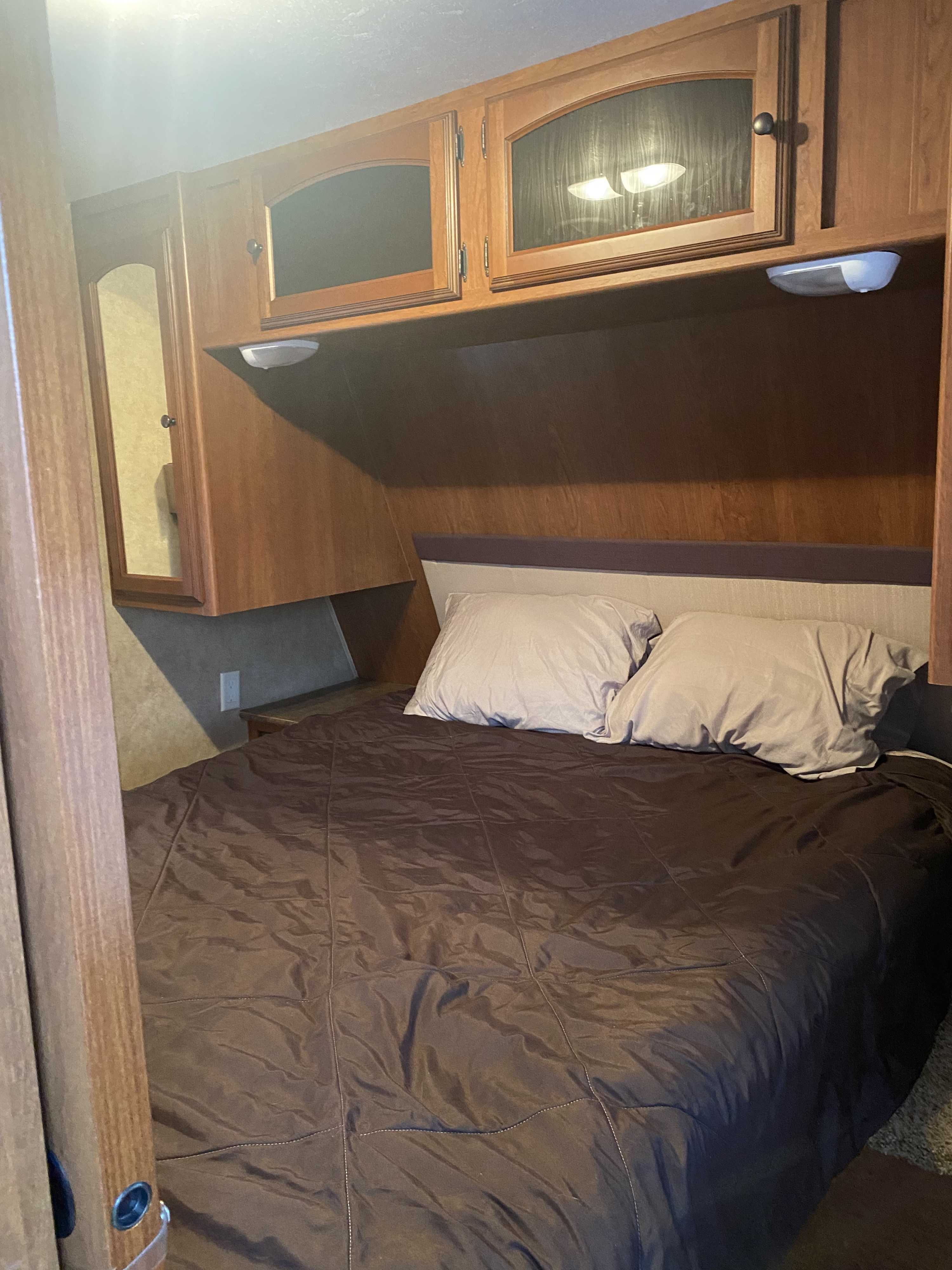 Cozy RV rental bedroom features a bed with brown comforter, light pillows, wooden cabinets with glass fronts, and under-cabinet lights.. Dutchmen Dutchmen Trailer 2013