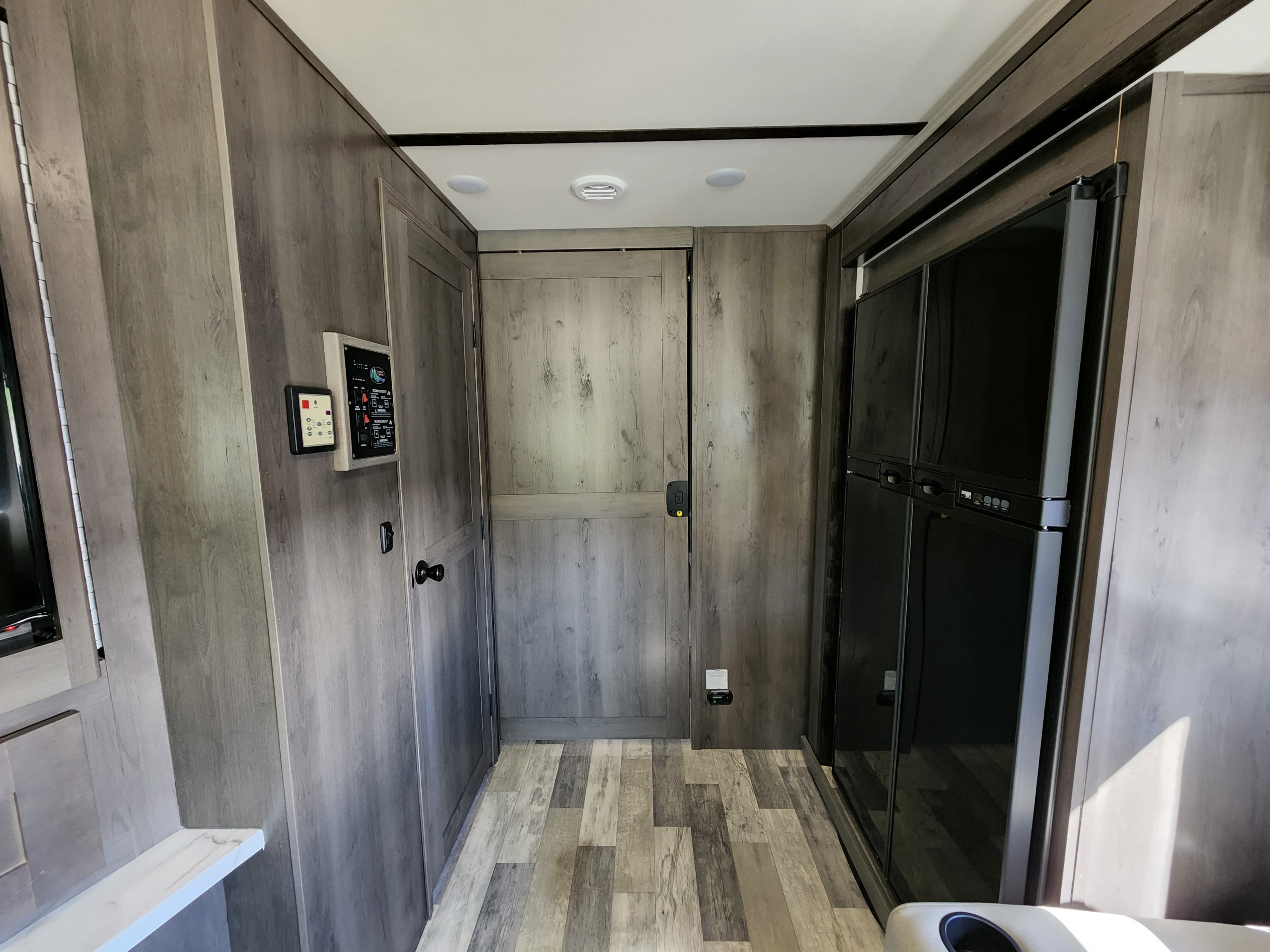 Modern RV rental interior with wood-grain walls/floor, control panels, closed door, and large black refrigerator.. Forest River FR3 34DS 2022