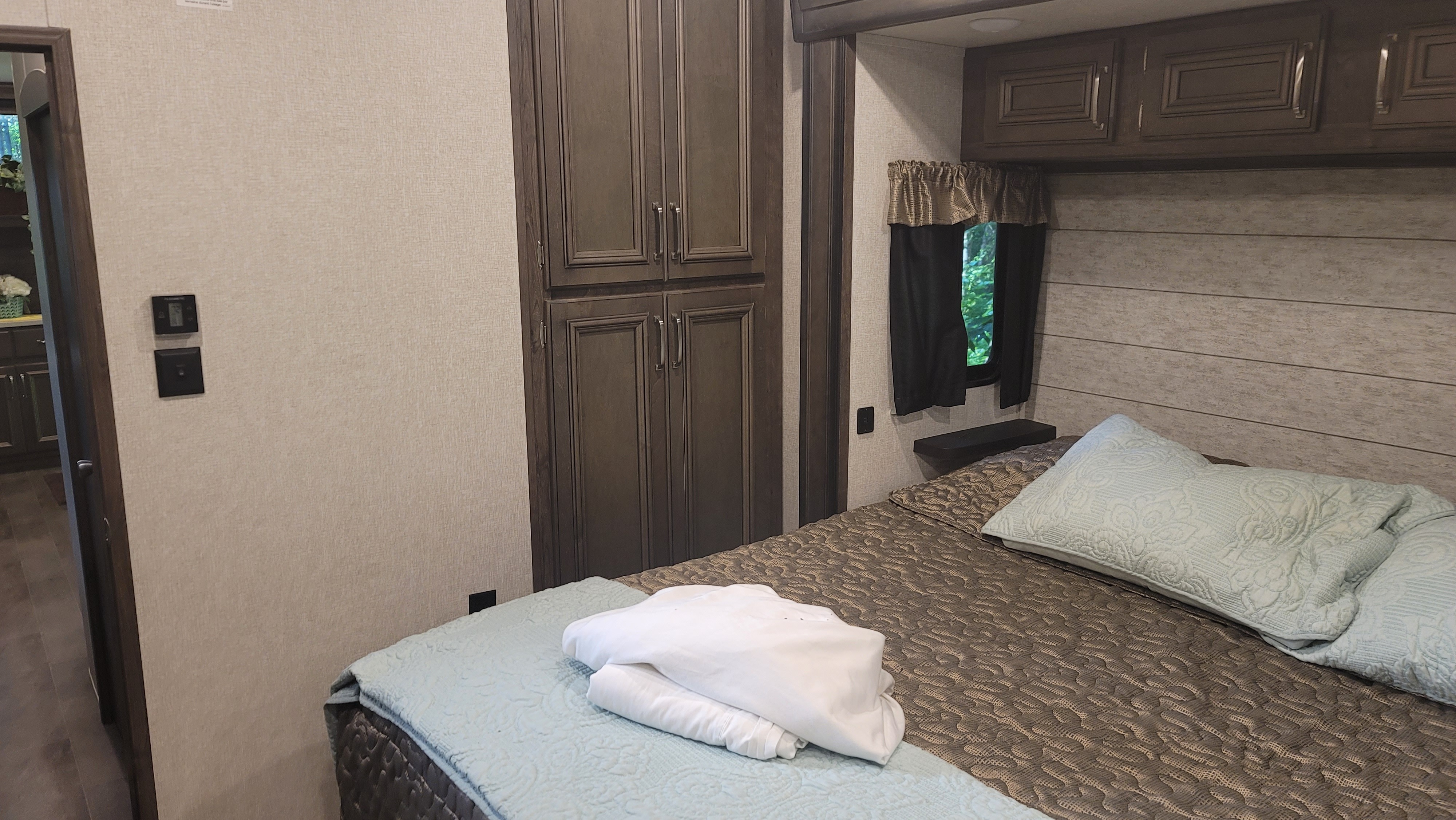 This comfortable RV rental bedroom features a cozy bed with blue/brown bedding, dark wood cabinets, and a window. Ready for your trip!. Keystone RV Montana High Country 2019