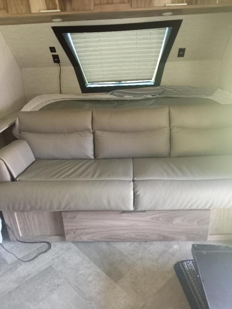 RV rental interior: A beige sofa sits before a bed with an angled window and blinds, plus wood overhead cabinets.. Forest River Palomino Palomini 2022