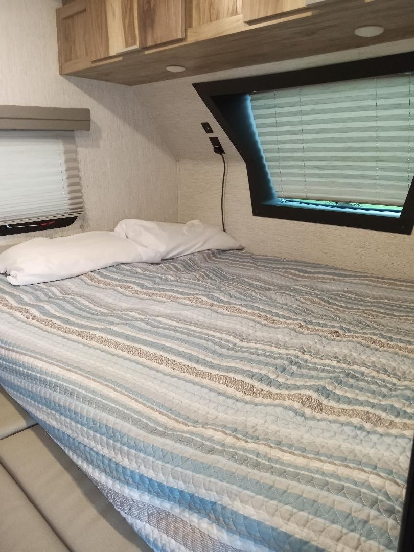 An RV rental bedroom showcasing a comfortable bed with striped comforter, white pillows, overhead cabinets, and two windows with blinds.. Forest River Palomino Palomini 2022