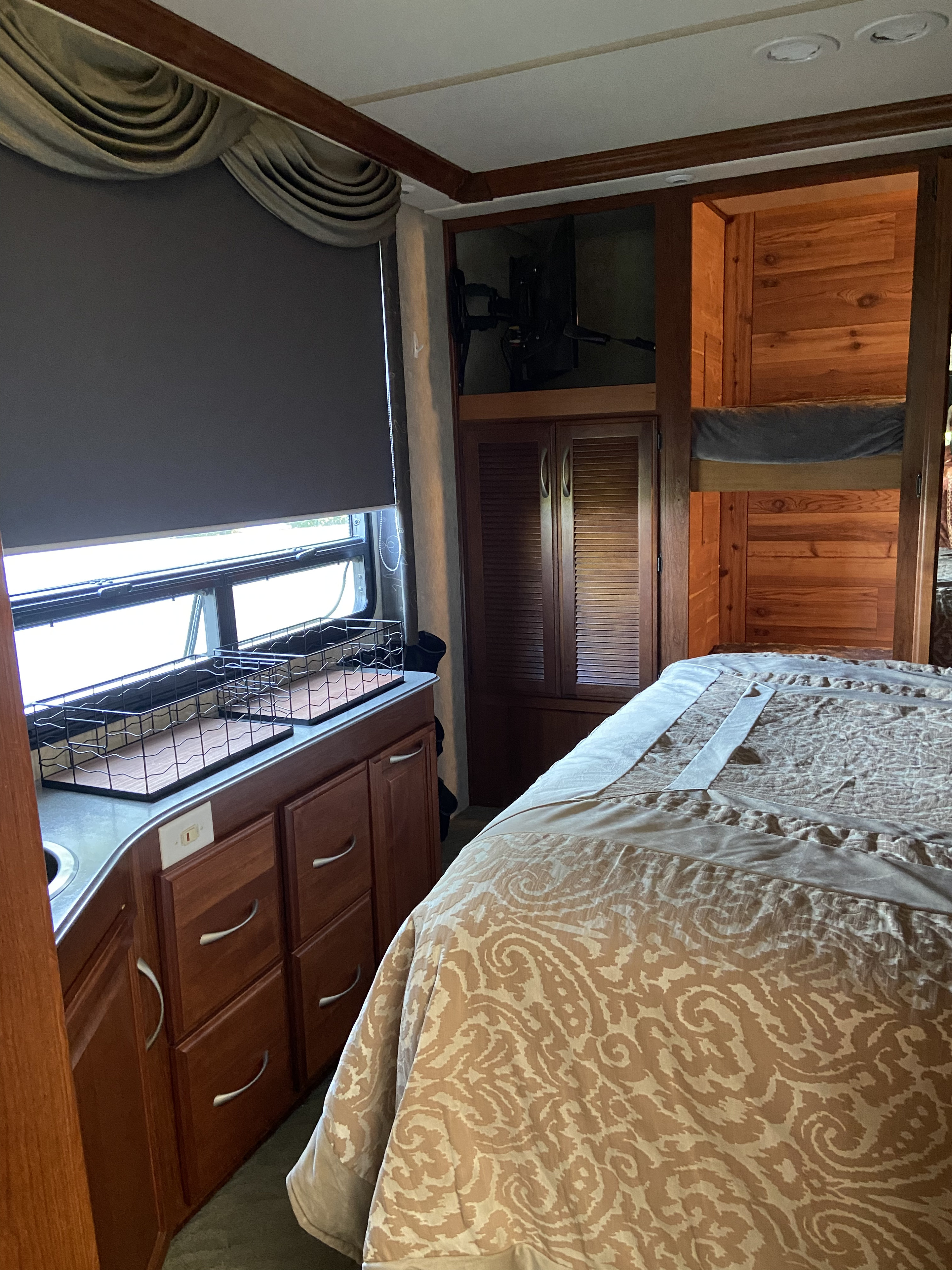 Inside an RV rental: bedroom area with a patterned bed, wood cabinetry, small counter with sink, and window with shade.. Fleetwood Providence 2005