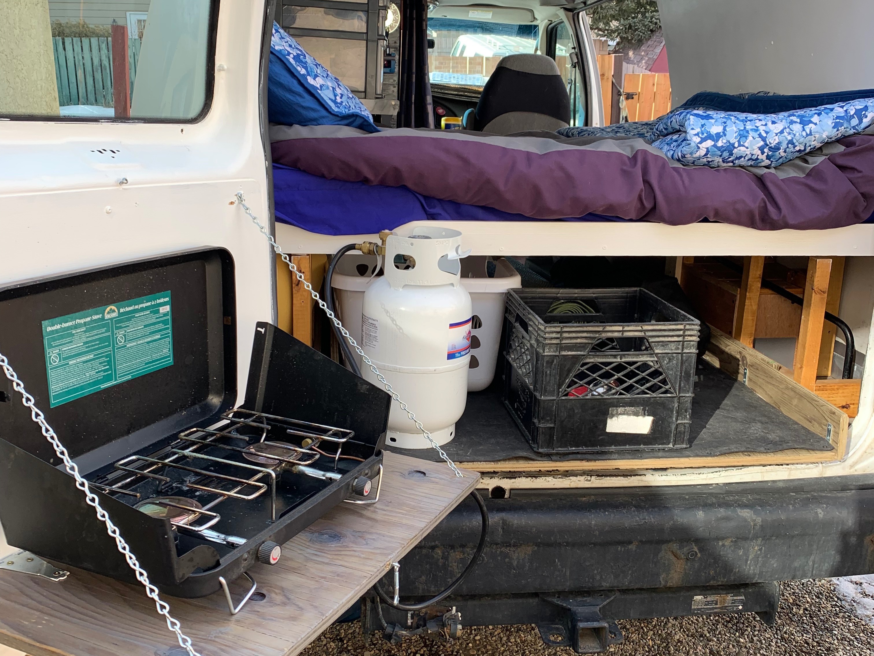 RV rental: Camper van's side door open revealing an outdoor propane stove on a shelf, and inside, a bed with storage.. Ford E350 Box Van 2005