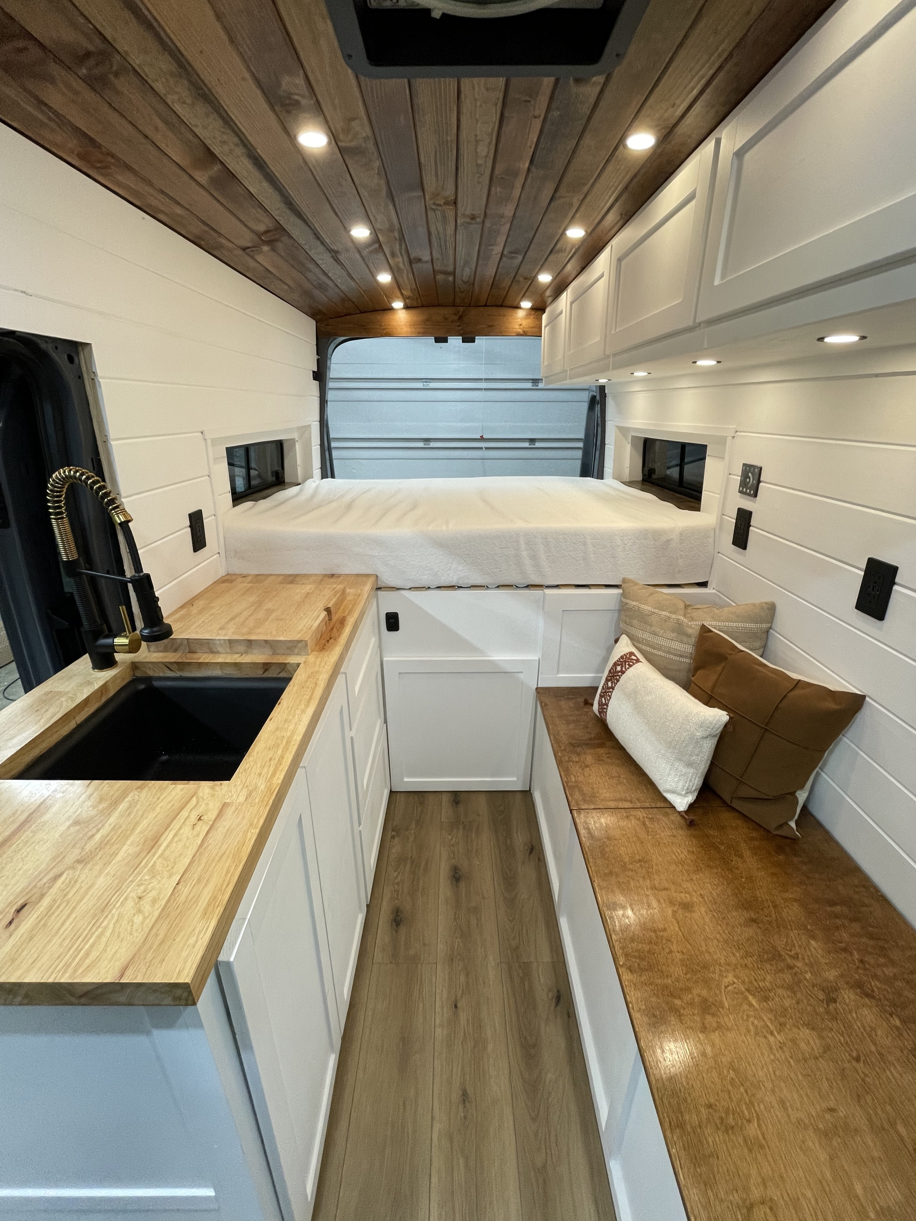 Step inside this RV rental! Cozy camper van interior featuring a kitchen, bed, and bench seating with wood details.. Ford Transit 2021