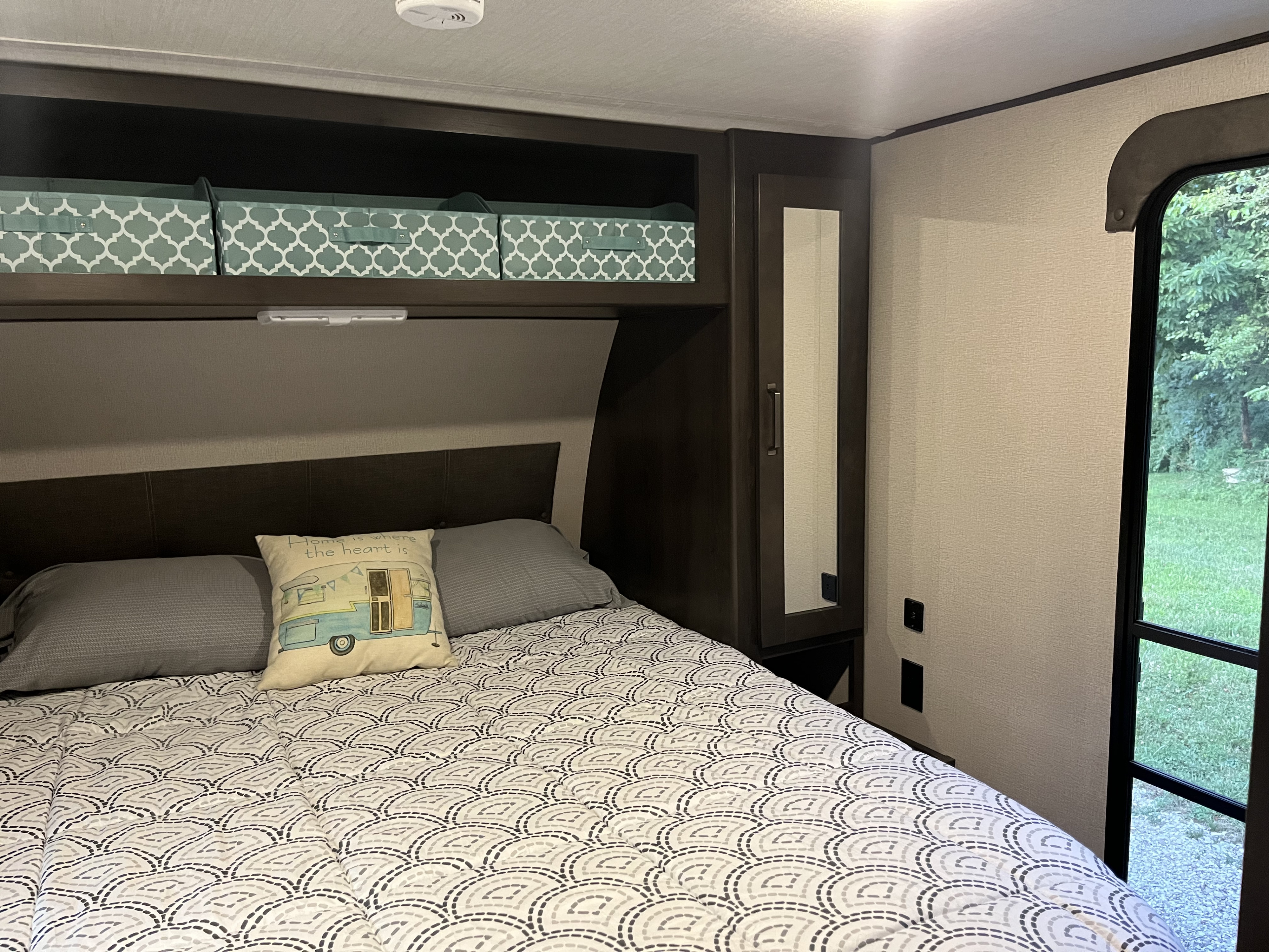 Cozy RV rental bedroom with patterned bedding, overhead bins, and a mirrored wardrobe, offering a view of greenery.. Grand Design Transcend Xplor 2020
