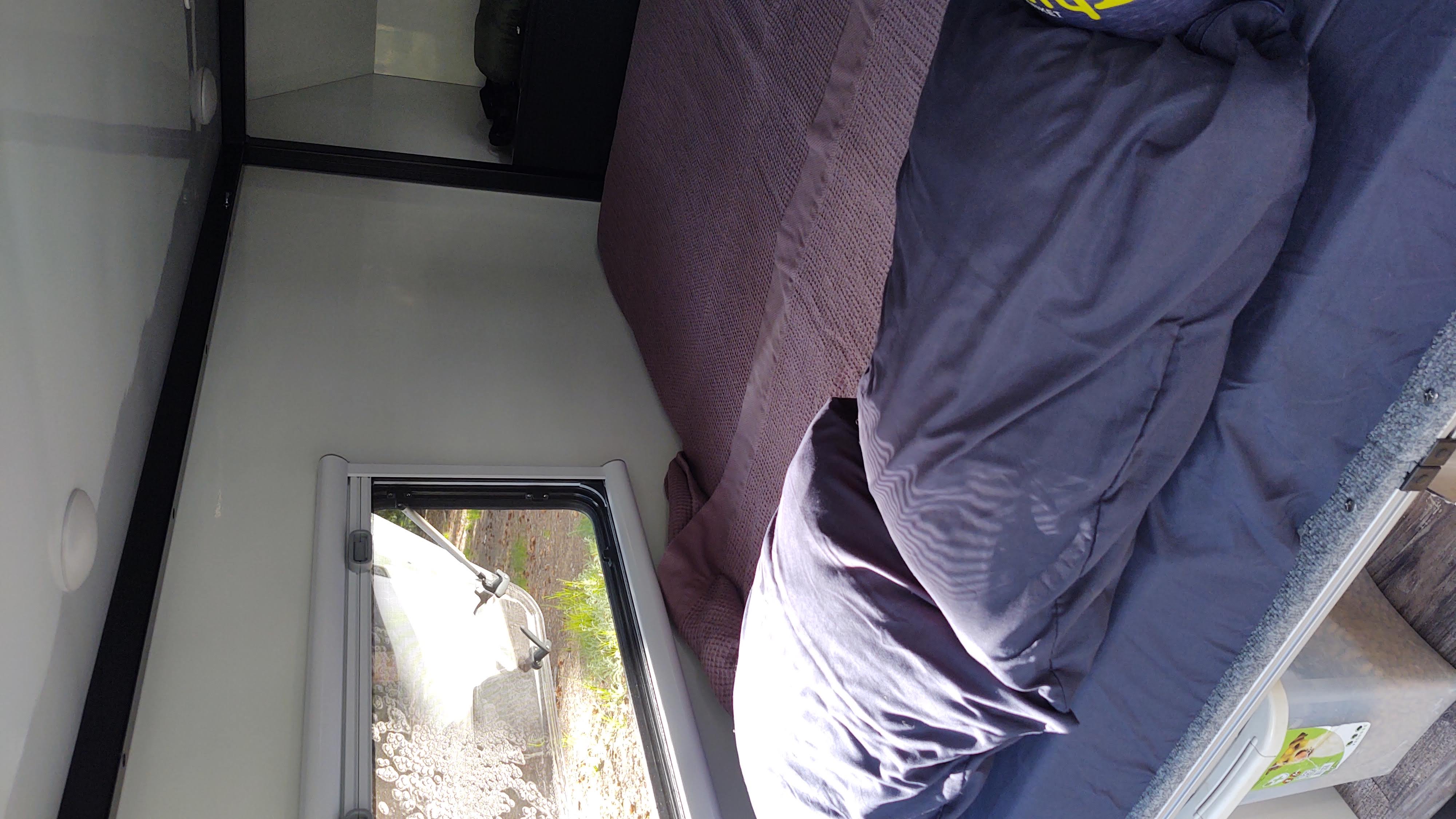 Inside this RV rental: A cozy sleeping space with a bed, blue bedding, purple blanket, window, and floor storage.. TetonX Porter 2022