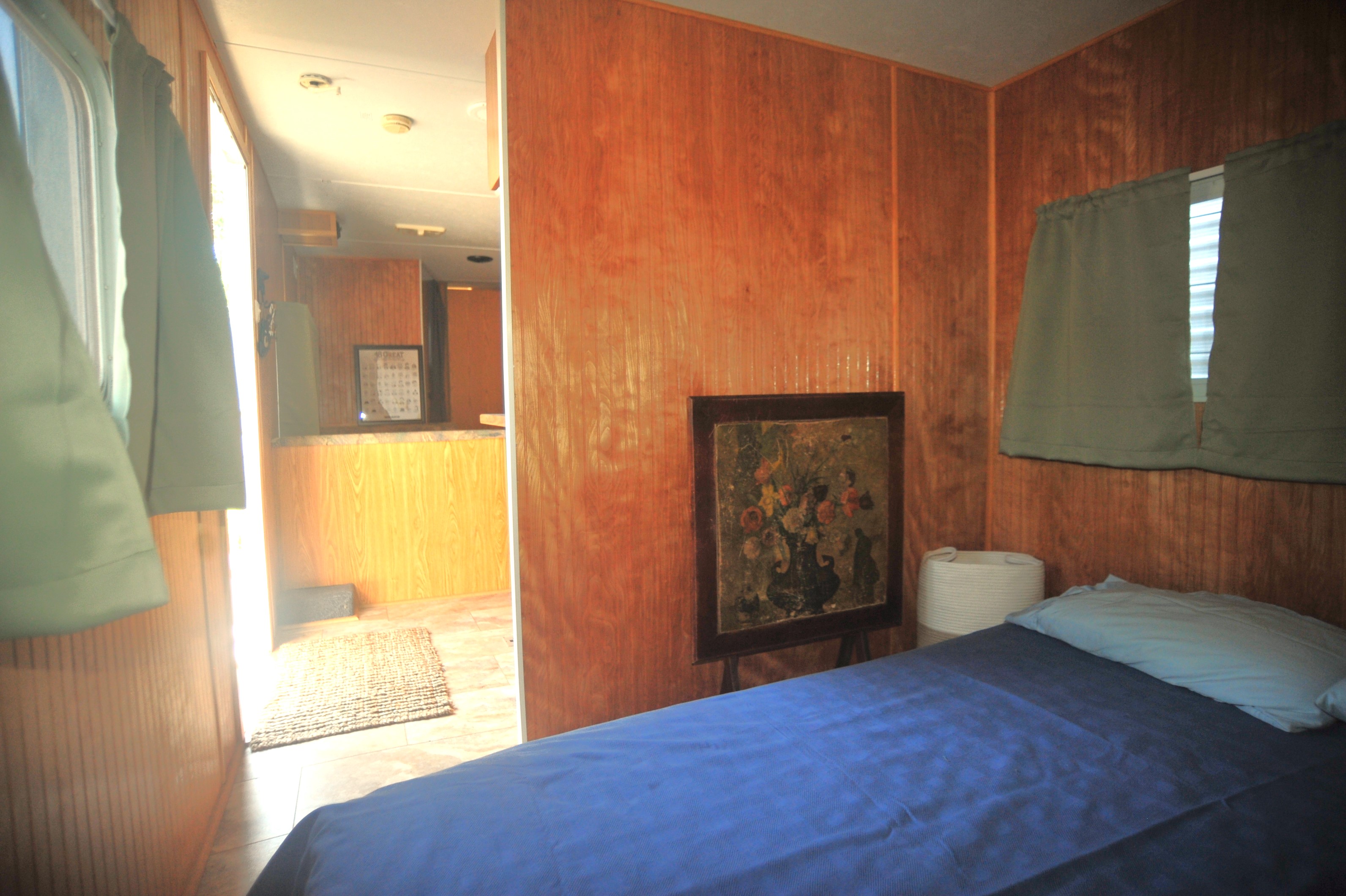 RV rental interior: a compact bedroom with wood-paneled walls, blue bed, floral painting, and doorway to another area.. Dutchmen Sport 2002