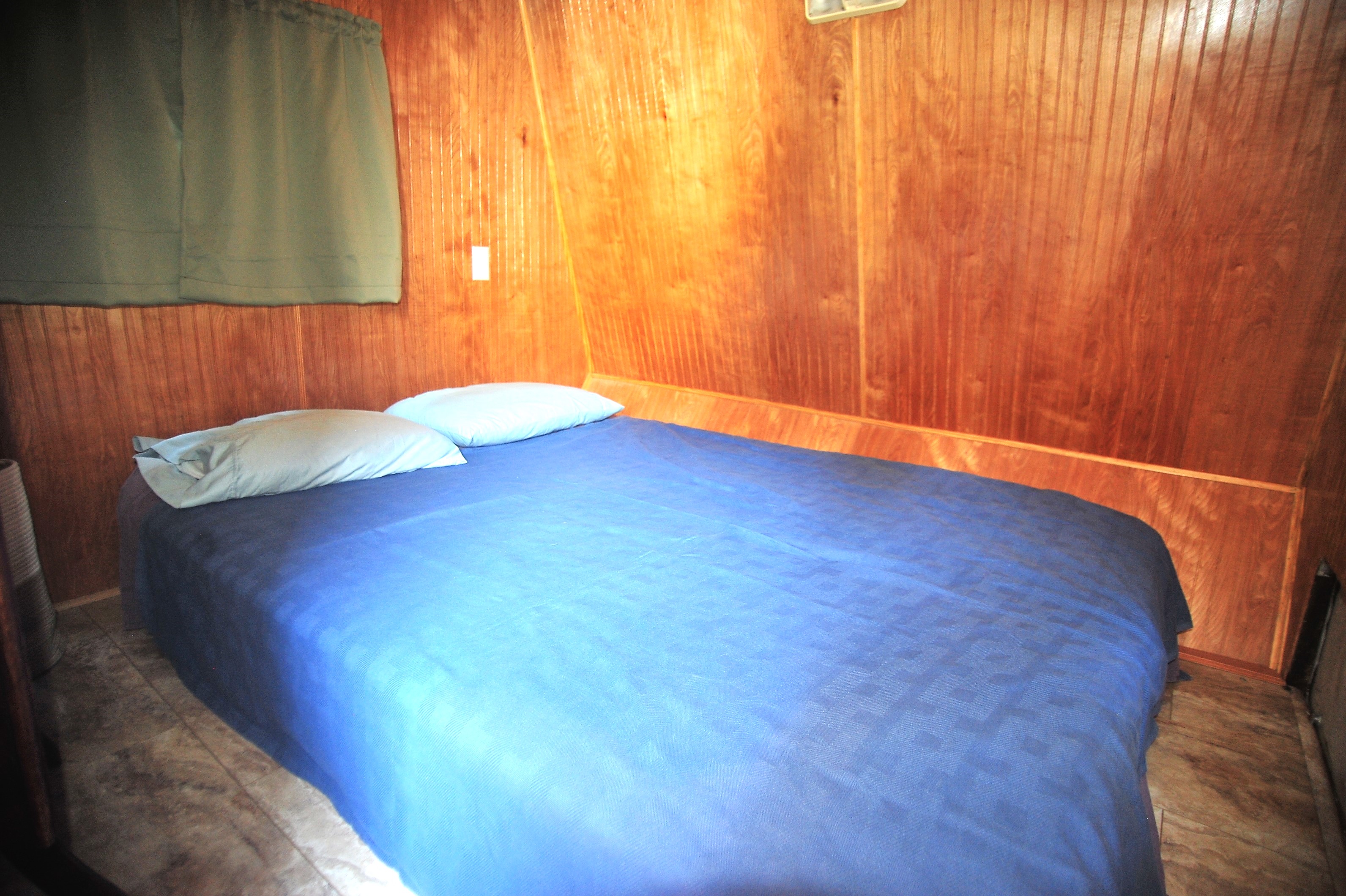 RV rental bedroom featuring a bed with blue bedding, light blue pillows, warm wood paneling, and a green curtain.. Dutchmen Sport 2002