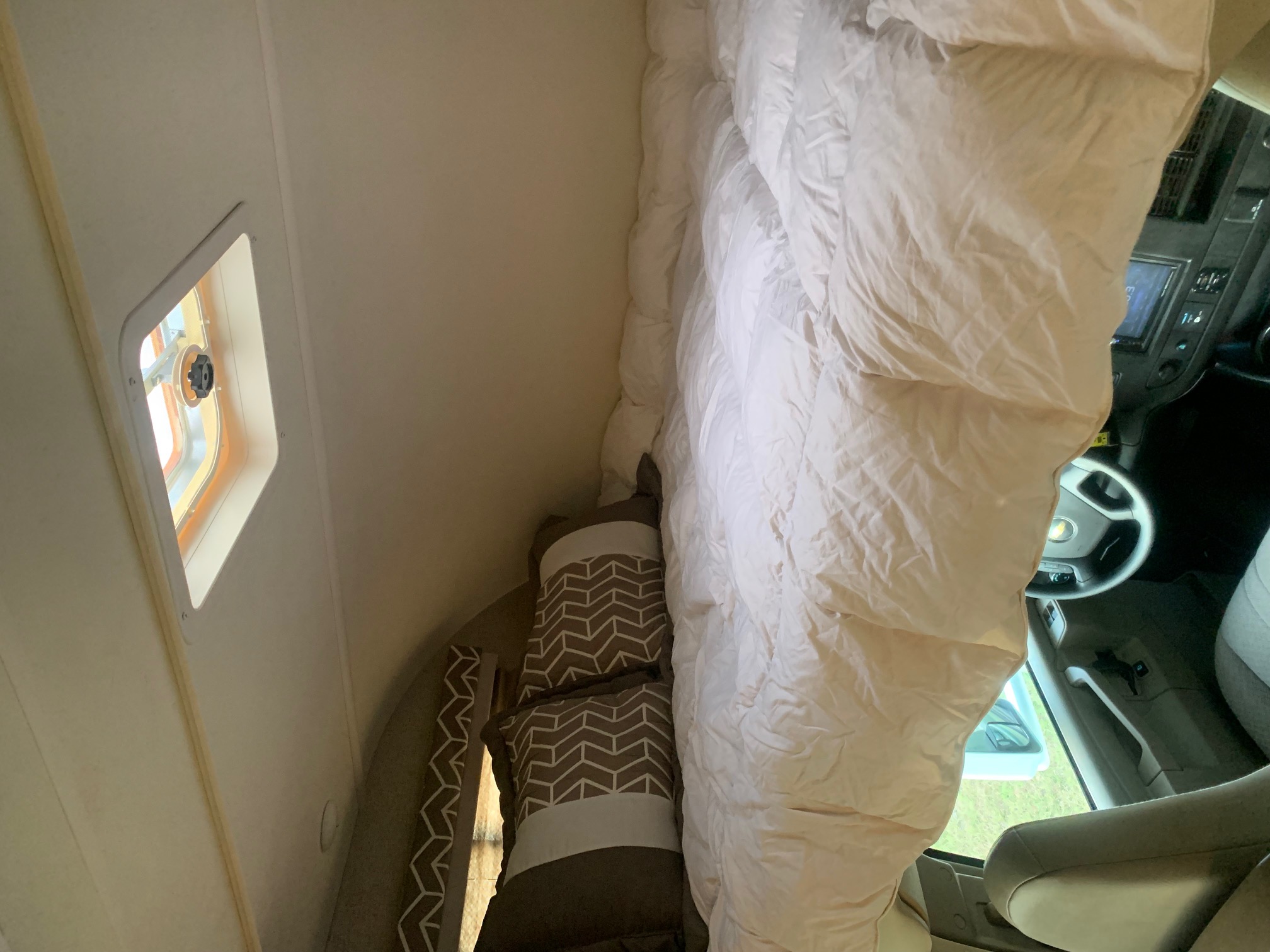 Bright RV rental interior: overhead bunk with white comforter, patterned pillows, a small window, and the cab.. Thor Daybreak 2020
