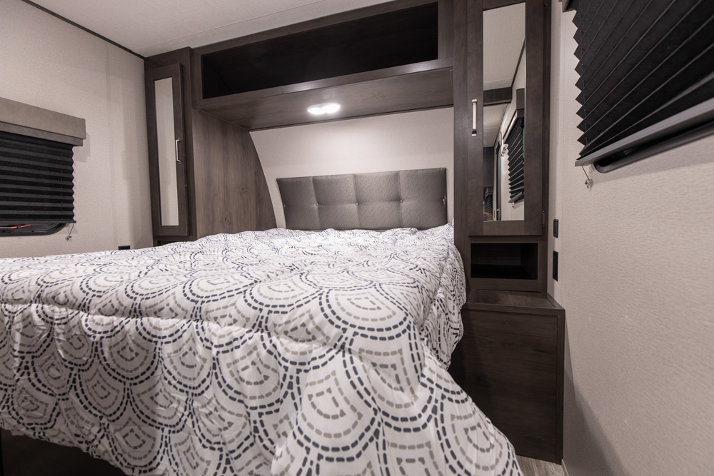 RV rental bedroom interior: a comfortable bed with a patterned comforter, padded headboard, overhead lighting, and compact storage.. Grand Design Transcend Xplor 2022