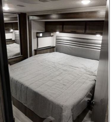RV rental bedroom: a comfortable bed with gray comforter, overhead storage, and mirrored wardrobe on the left.. COACHMEN LEPRECHAUN 2021