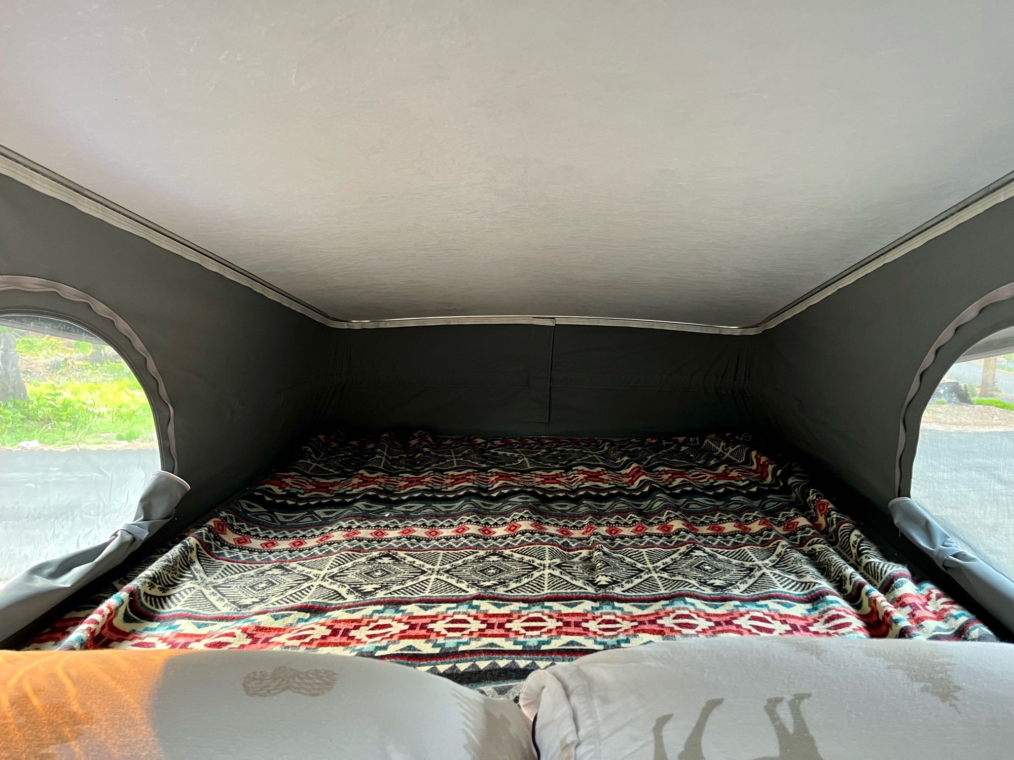 Cozy RV rental pop-top bed with vibrant patterned blanket. Windows show lush greenery outside.. Winnebago Solis 2021