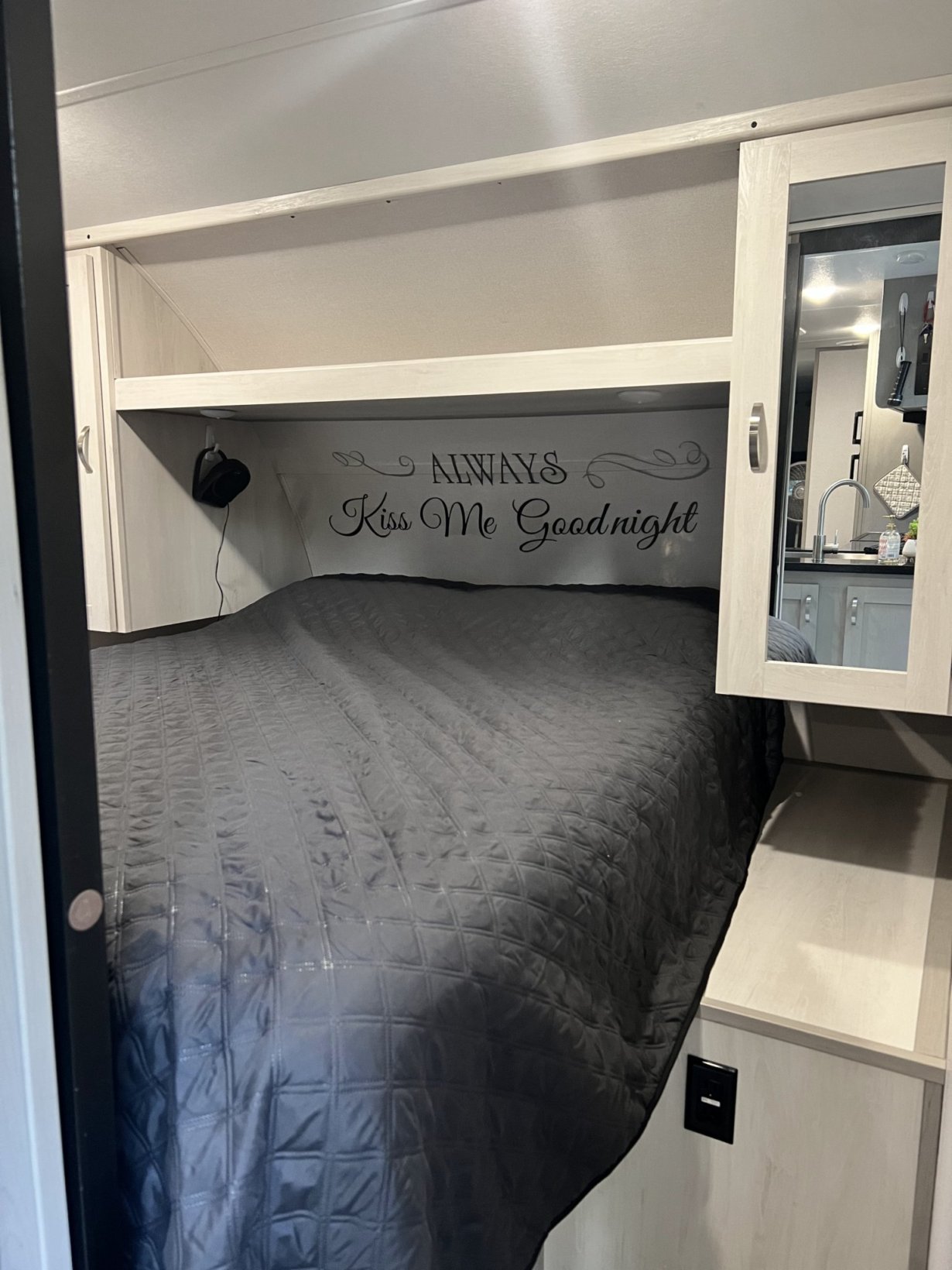 Compact RV rental bedroom with dark grey bed, shelving, and a "Kiss Me Goodnight" decal. Mirror reflects the kitchen.. PrimeTime 31BHD 2021