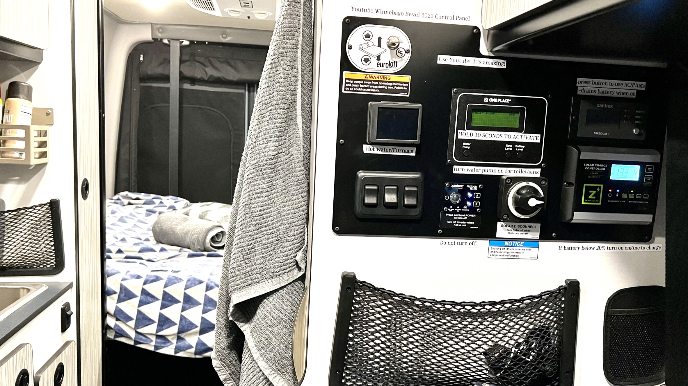RV rental interior: A camper van's control panel, showing switches and displays for heating, water, and solar charge, beside a cozy bed.. Winnebago Revel 2022
