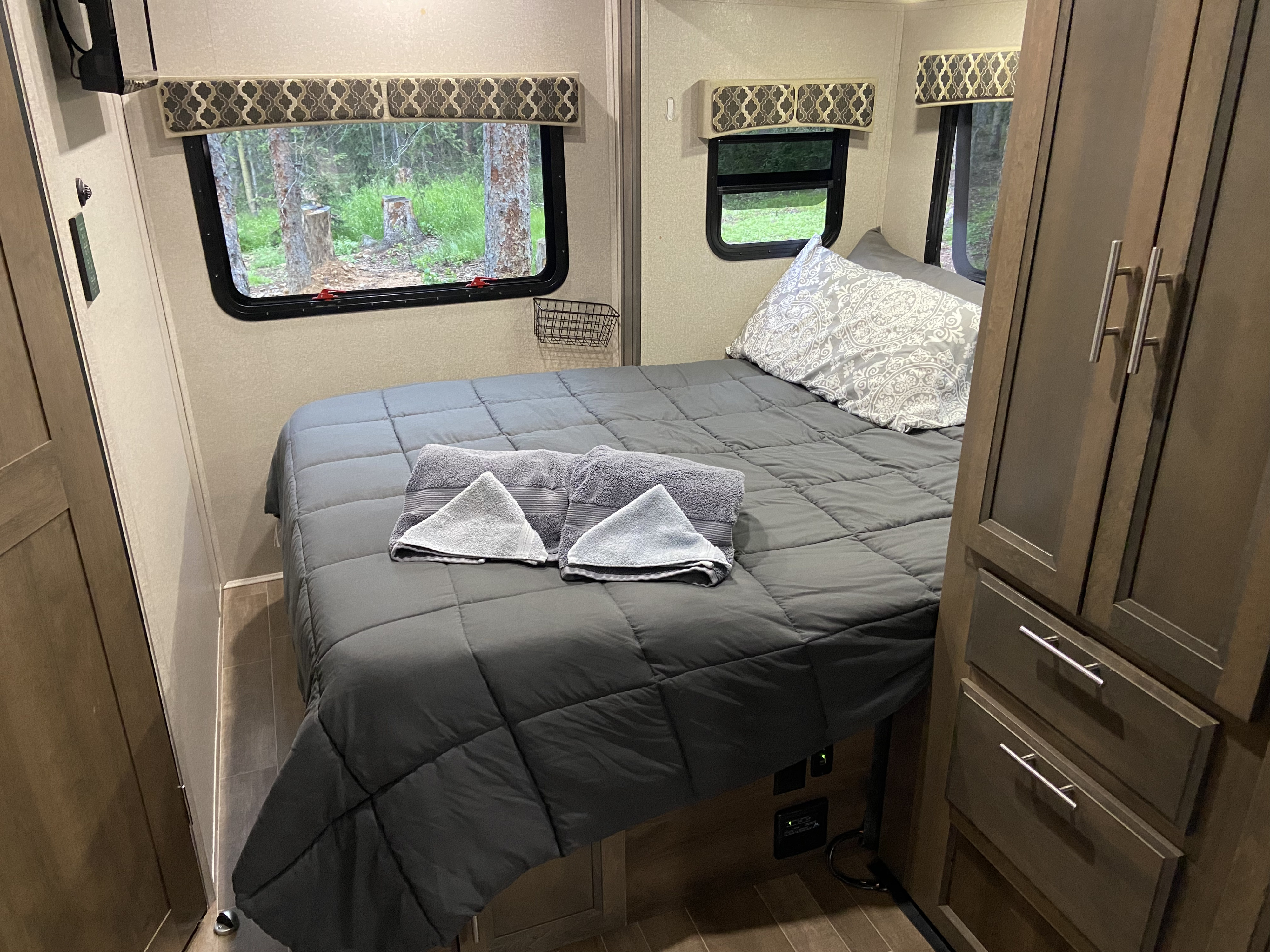 Cozy RV rental bedroom features a comfortable bed, built-in storage, and serene nature views from large windows.. Dynamax ISATA 3 2020
