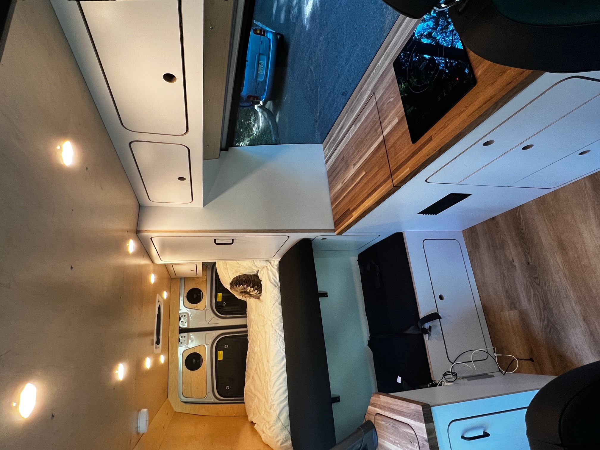 Modern RV rental interior with cozy bed, kitchen cooktop, custom storage, and view of a car on the road outside.. VanCraft Mercedes Benz 2022