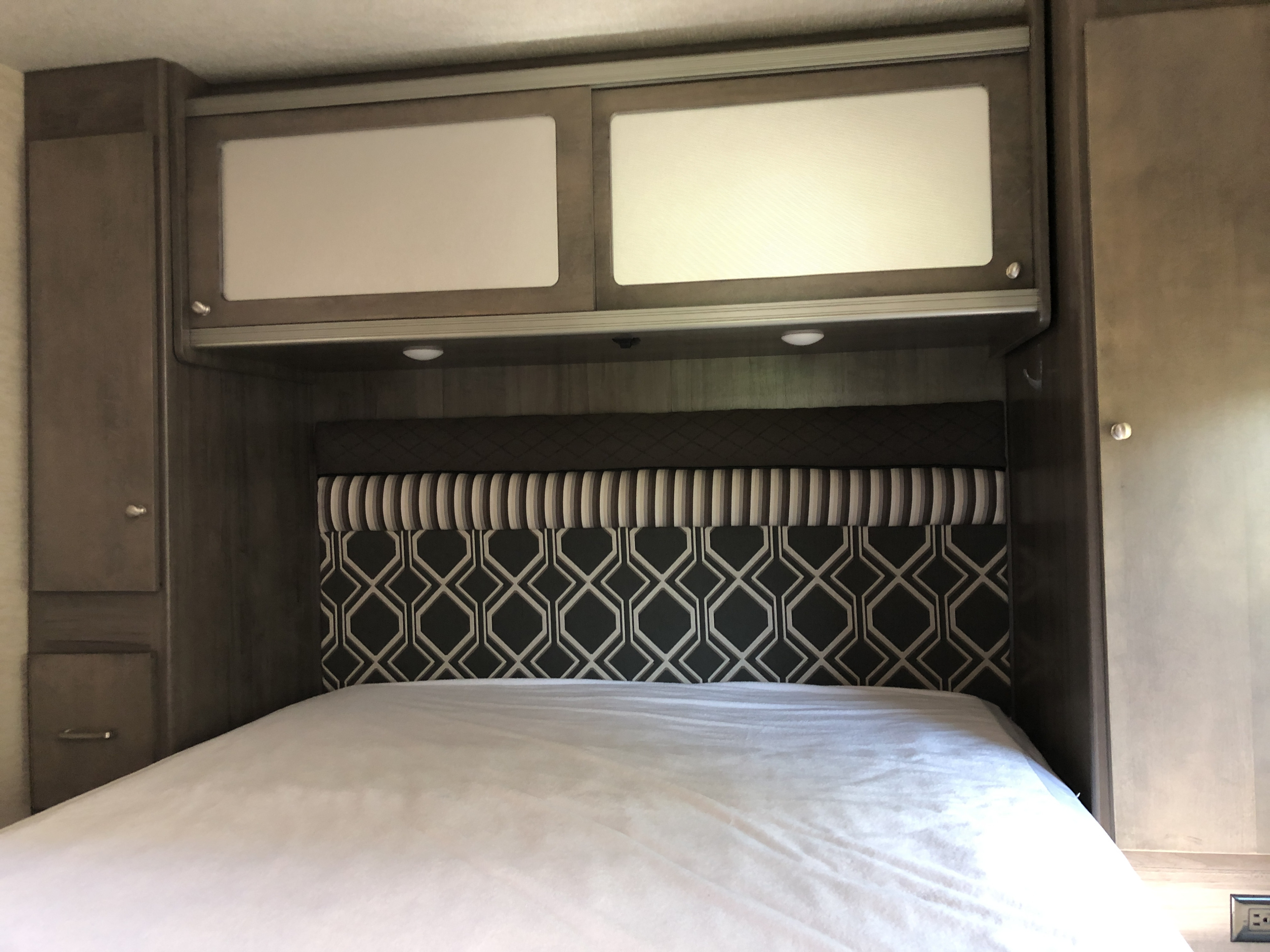 RV rental bedroom: Compact interior with a patterned headboard, overhead sliding cabinets, & convenient lighting.. Winnebago Brave 2016