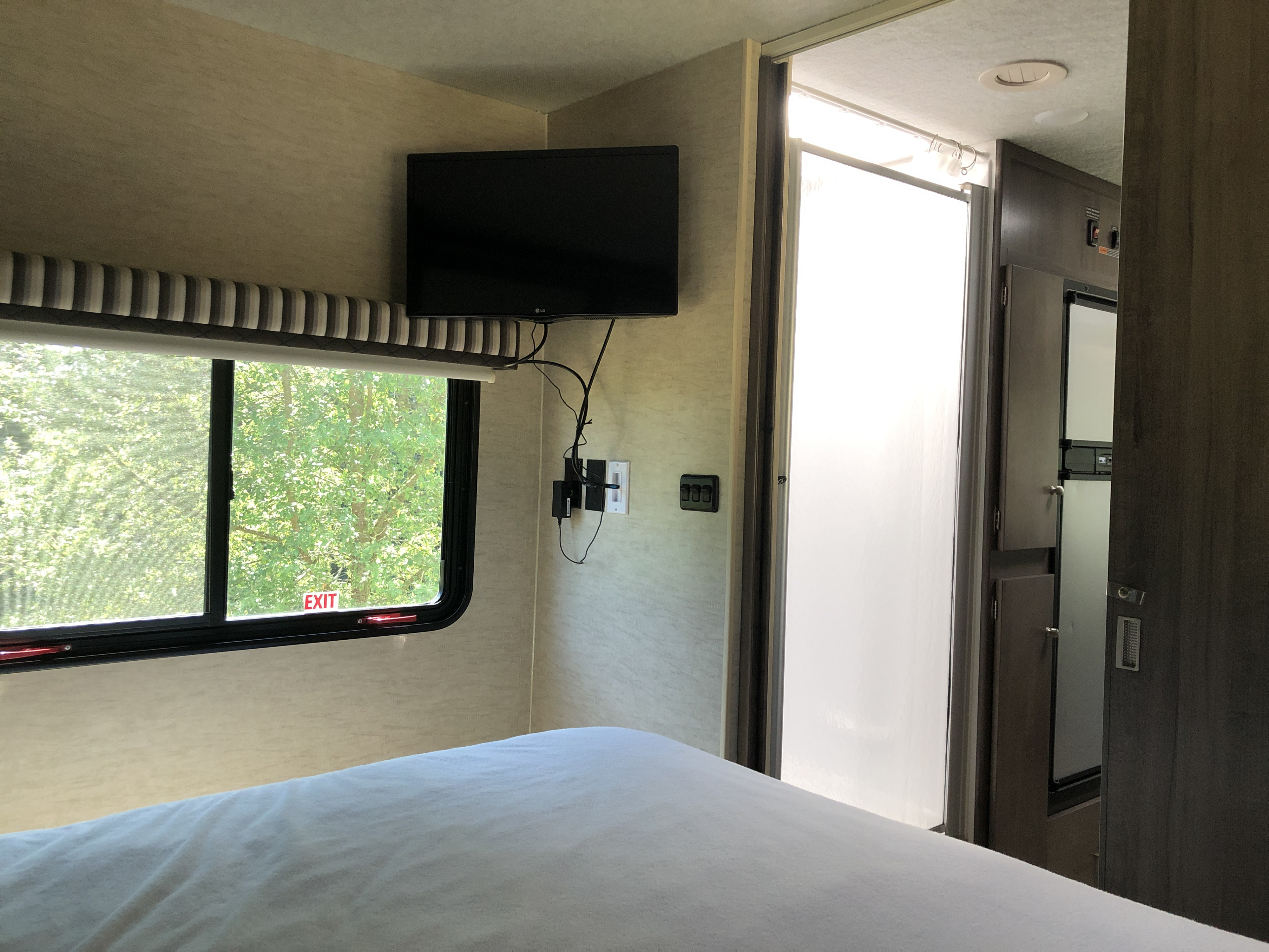 Explore this RV rental's bedroom: TV, large window with "EXIT" sign, tree view, and door to the main cabin. White bed.. Winnebago Brave 2016